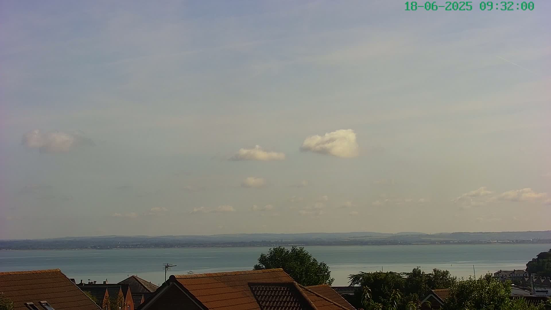 A partly cloudy sky overlooks a calm body of water and a row of brown-roofed houses.