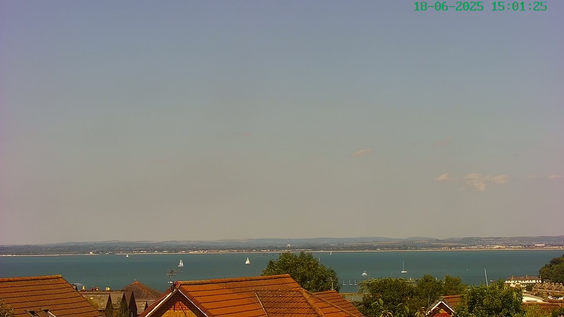 A hazy, sunny day overlooks a calm body of water with several sailboats and distant land, seen from behind the brown tiled roofs of houses.