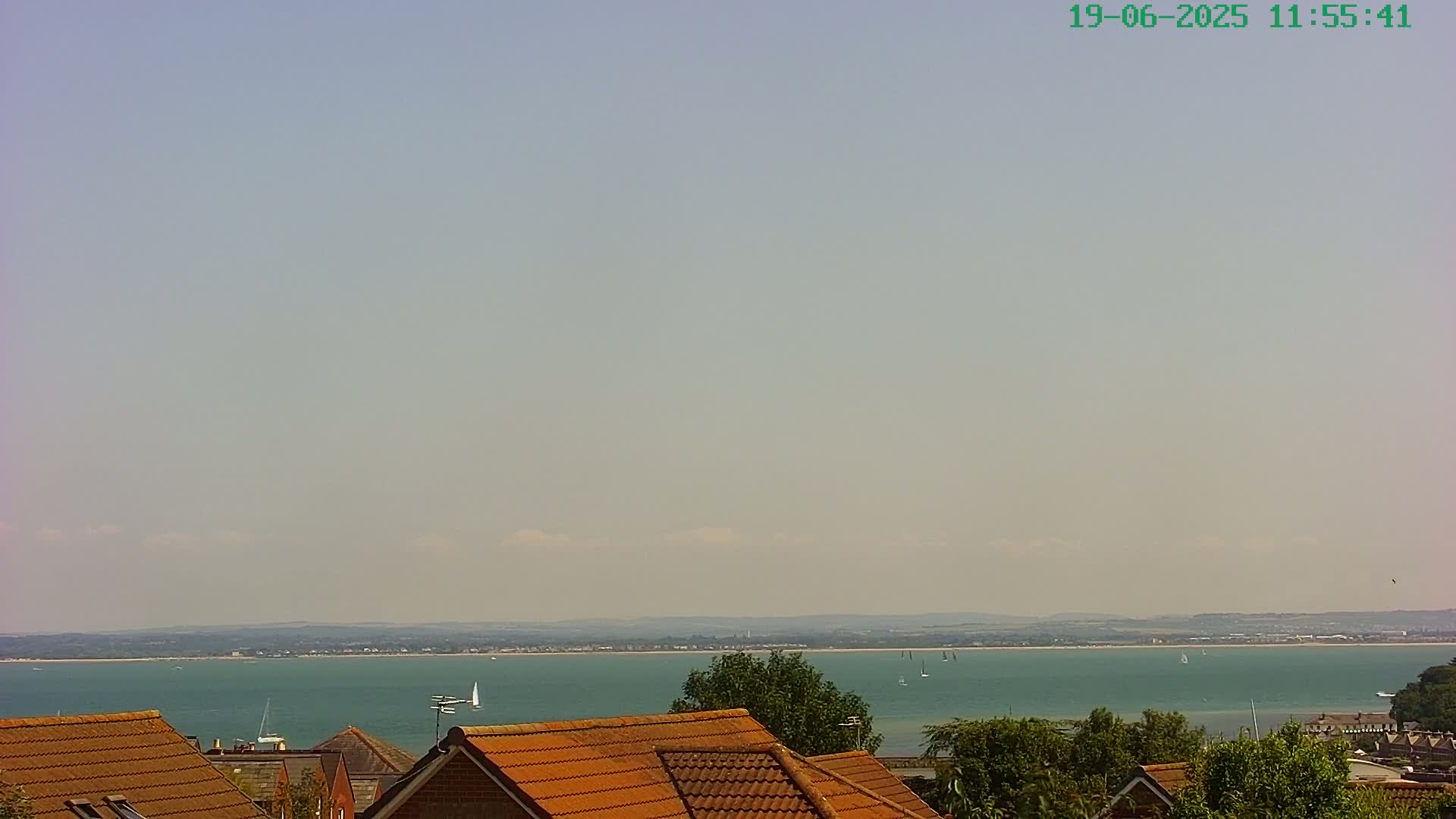 A mostly clear sky overlooks a calm, teal sea with several sailboats, viewed from above the terracotta roofs of houses and some trees.