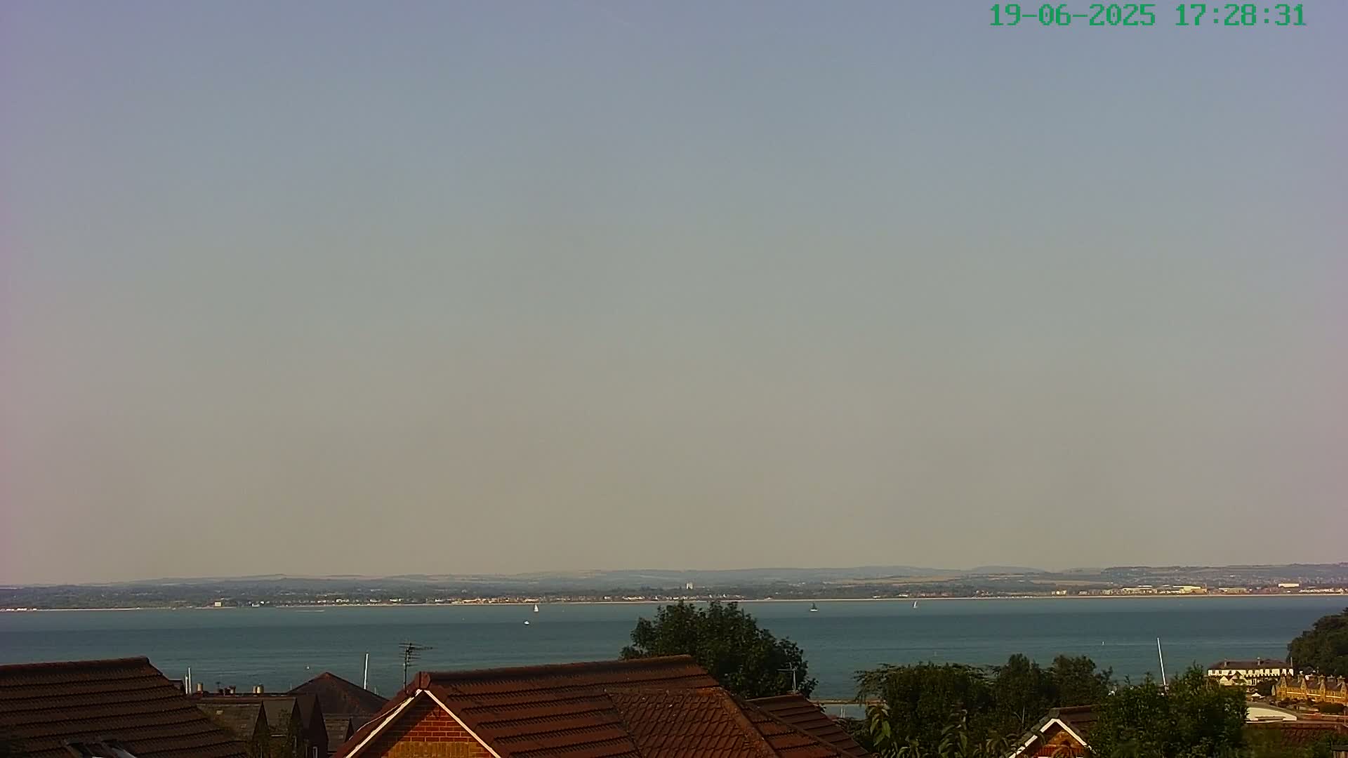 A hazy, calm day reveals a view of a calm body of water extending to a distant shoreline, with a few sailboats visible, seen from behind the rooftops of houses.