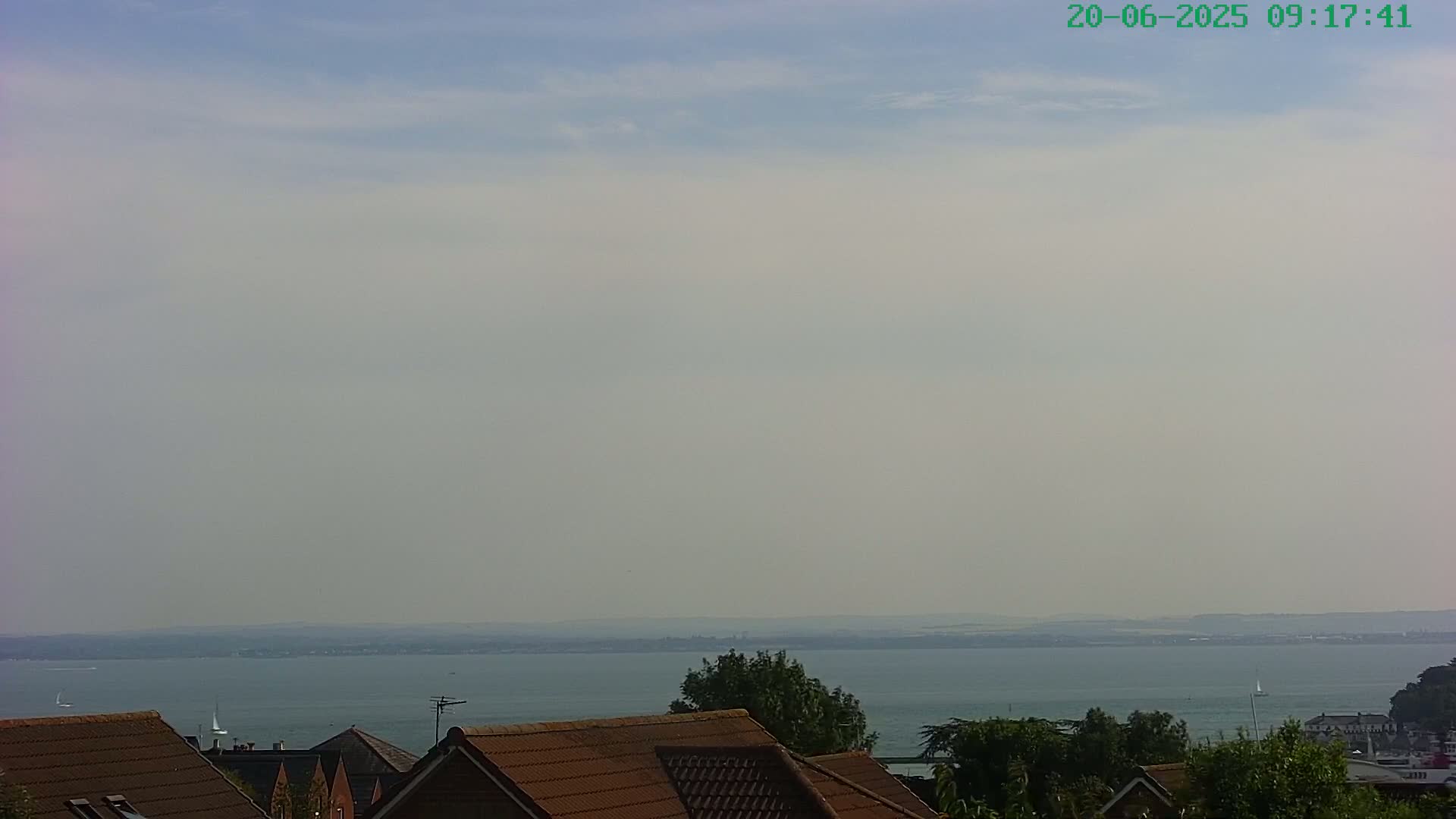 A hazy view of a calm body of water stretches to a distant shoreline, visible from behind the brown roofs of houses and some green trees.