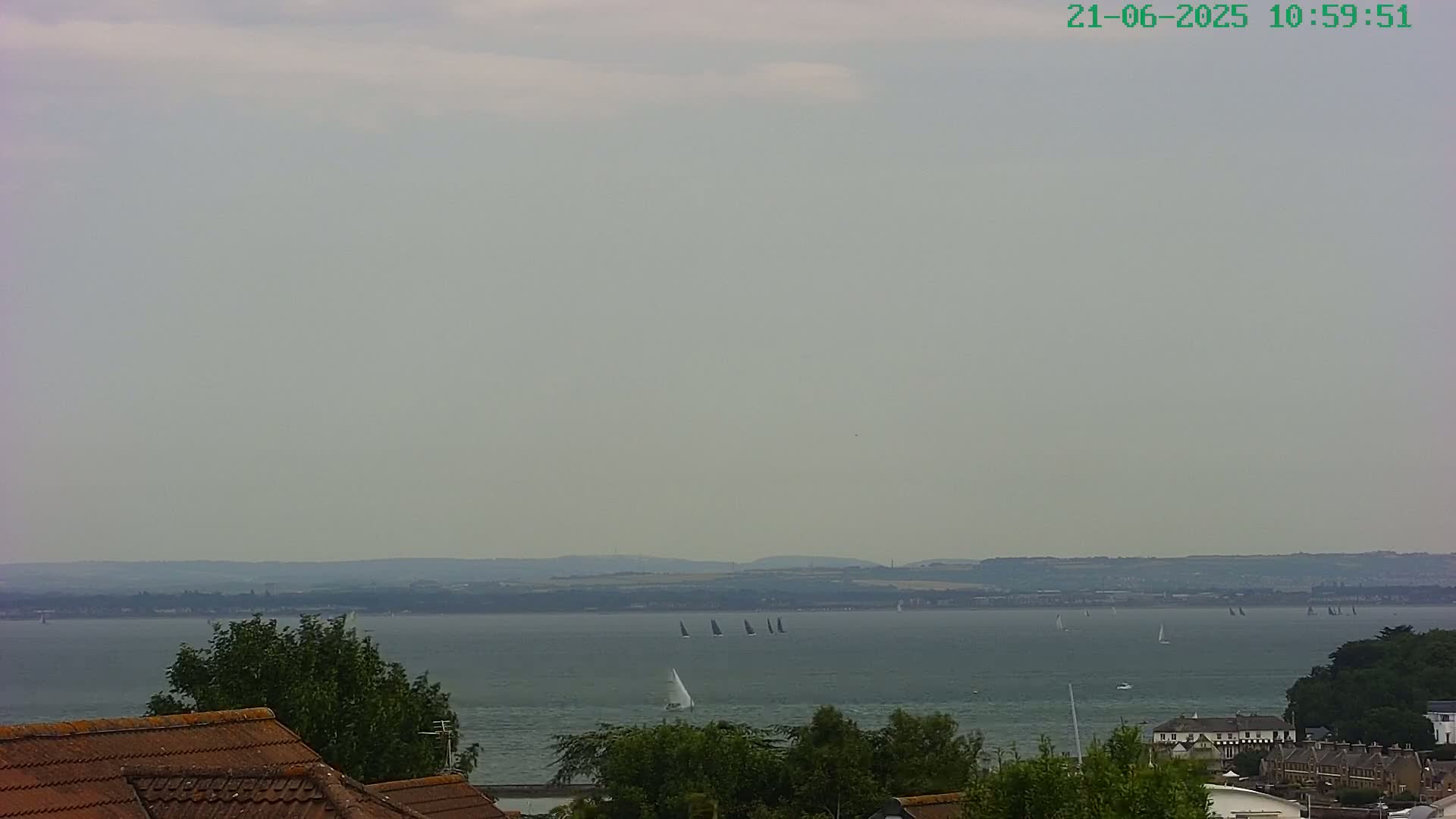 Several sailboats are visible on a calm, overcast body of water, with a distant shoreline and rooftops in the foreground.