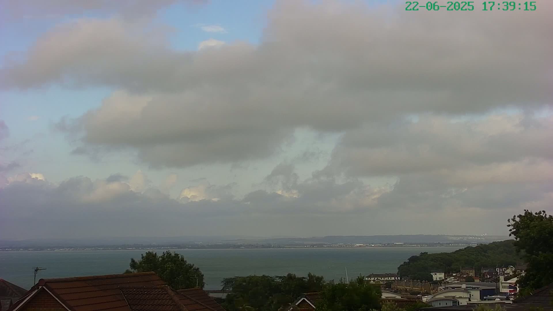 A mostly cloudy sky overlooks a calm body of water and a coastal town nestled among green trees.