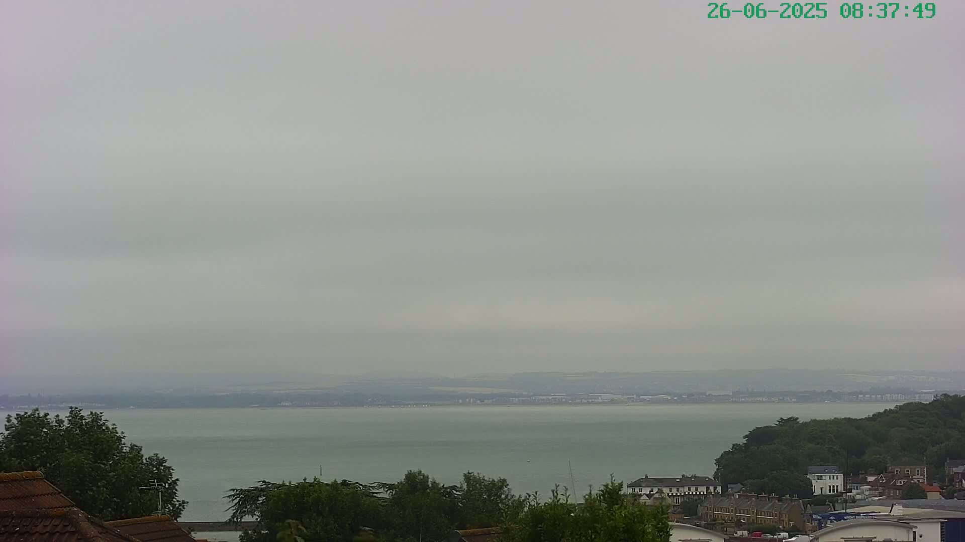 A calm, overcast day reveals a view of a wide body of water, bordered by land and a collection of buildings nestled among green trees.