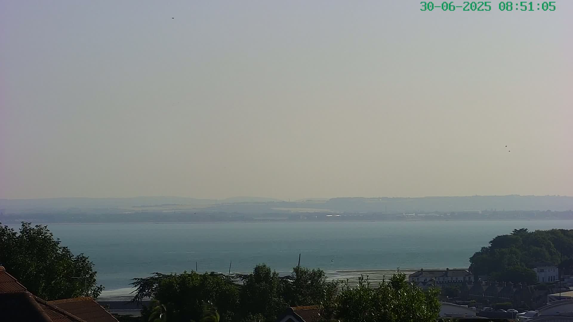 A hazy, calm day reveals a view of a large body of water, with distant land and a shoreline dotted with buildings and trees in the foreground.