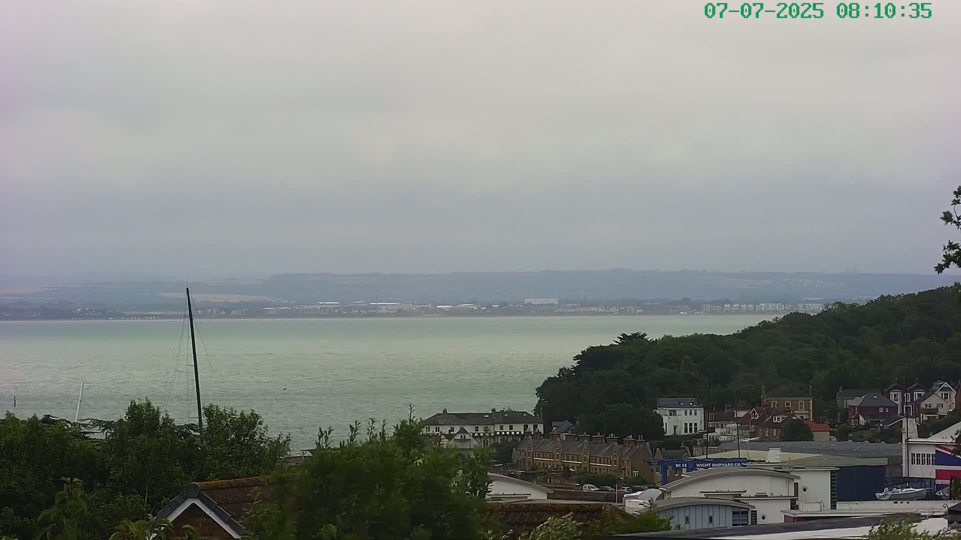 A view of a calm body of water under an overcast sky, with a shoreline featuring green trees and buildings in the foreground.