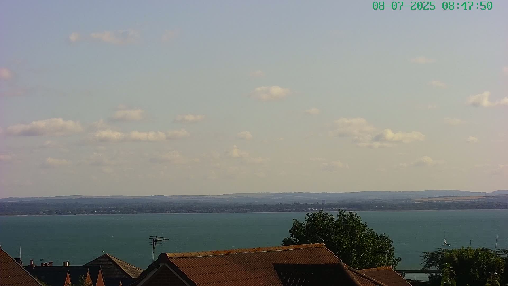 A partly cloudy sky overlooks a calm body of water and distant land, with the tops of several houses and trees visible in the foreground.