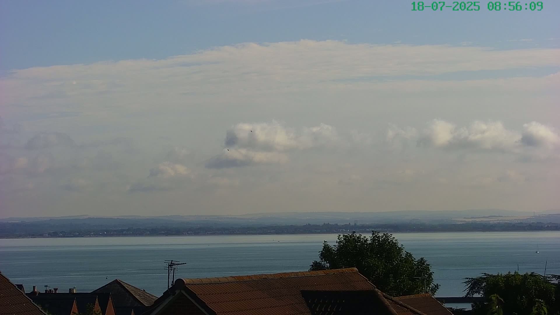 A mostly clear sky with scattered cumulus clouds overlooks a calm body of water and distant land, partially obscured by rooftops in the foreground.