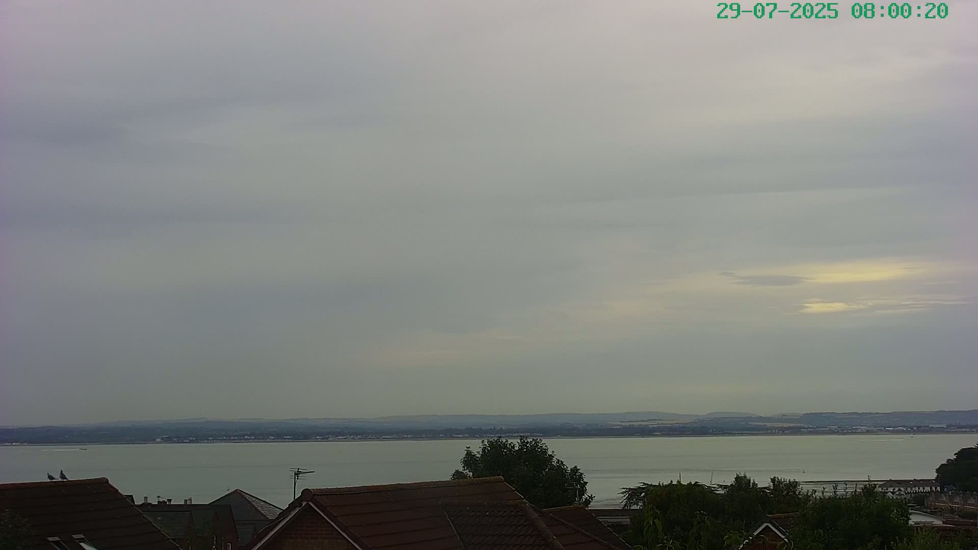 A calm body of water stretches to a distant shoreline under a cloudy sky, viewed from behind rooftops.