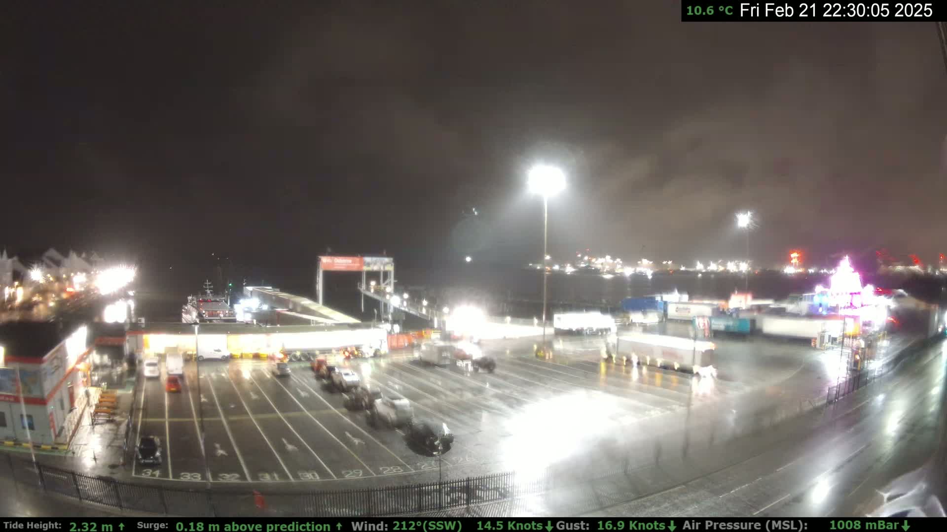 A nighttime view of a rain-soaked port area with vehicles in a parking lot, illuminated by bright lights, and ships visible in the background.
