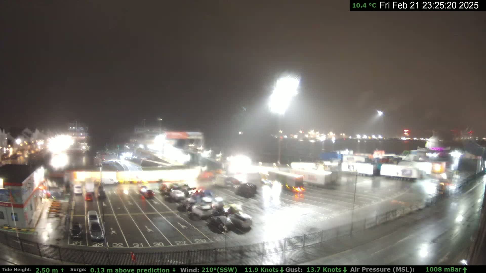 A nighttime view of a rain-soaked port with numerous vehicles parked in a lot near brightly lit buildings and large ships in the background.