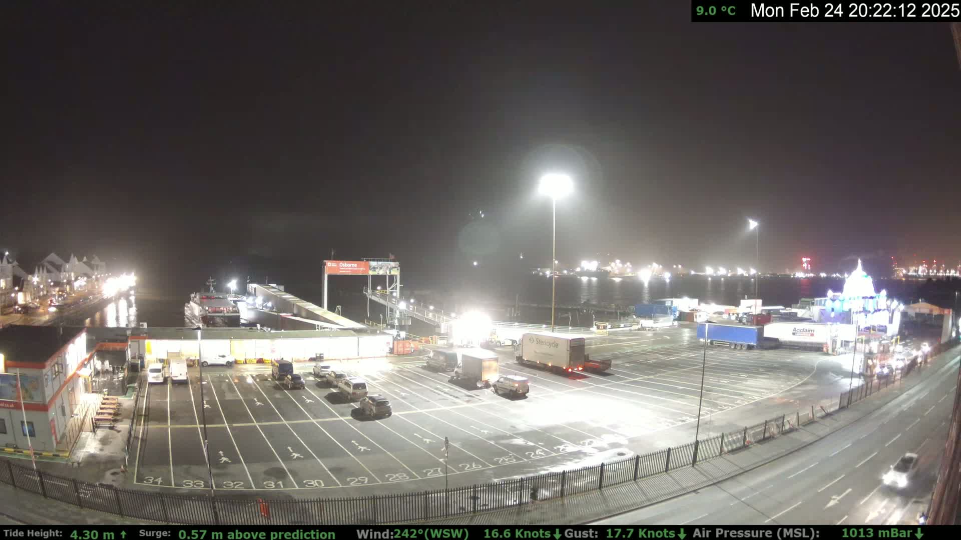 A nighttime view of a ferry terminal and adjacent parking lot with several vehicles, under a clear, dark sky.