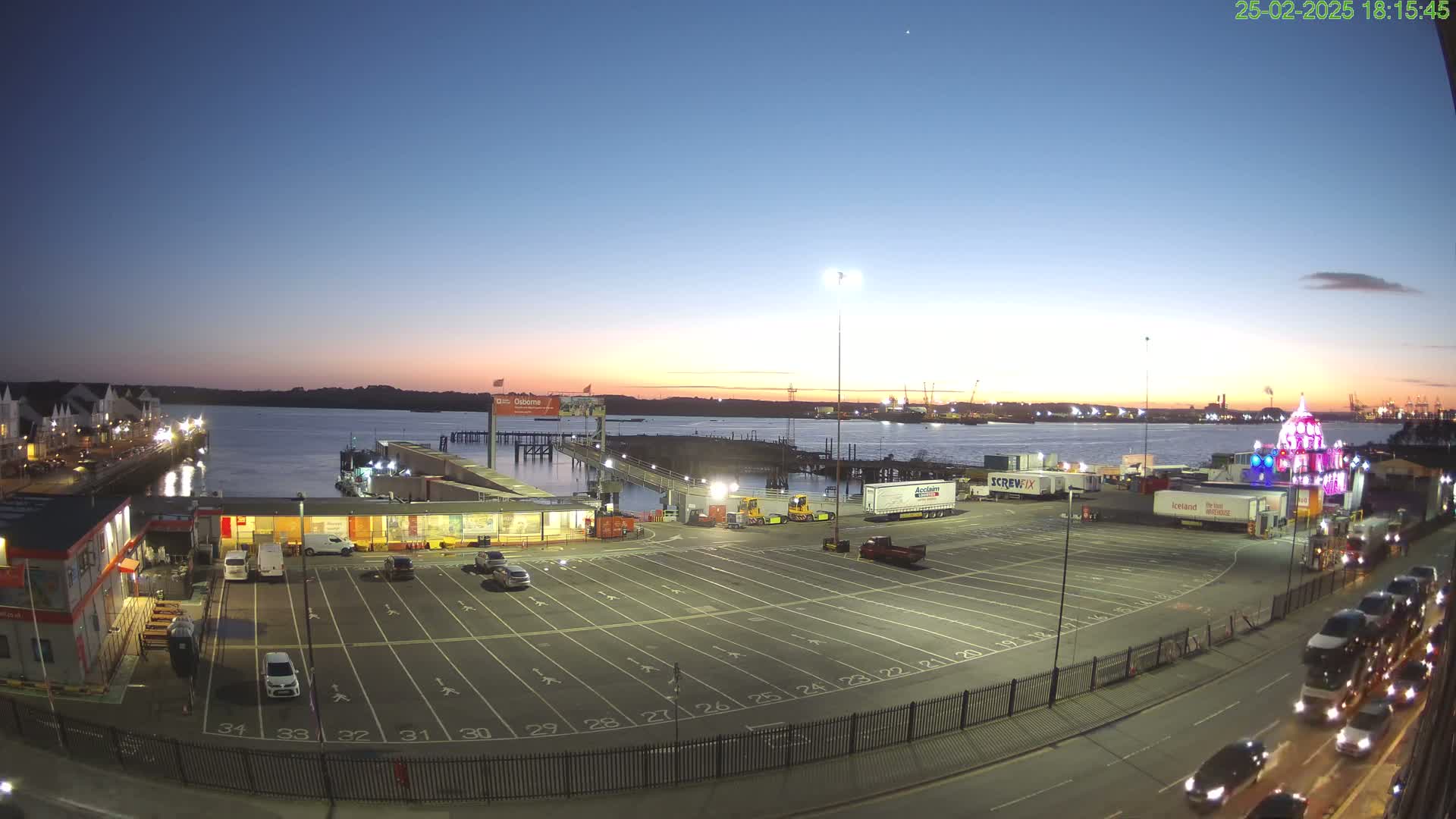 A twilight view of a ferry terminal with a large empty parking lot, a pier extending into calm water, and several vehicles parked near buildings.