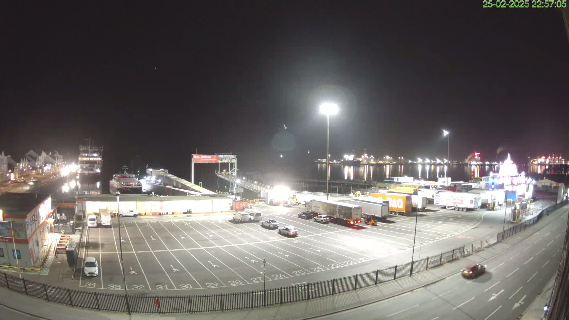 A nighttime view of a ferry terminal with several vehicles parked in a lot, ferries docked at the pier, and a calm body of water reflecting lights across the scene.