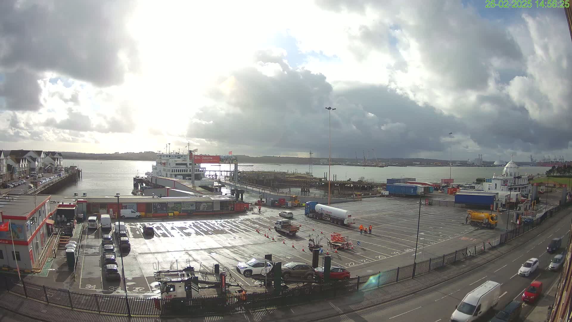 A ferry is docked at a port under a partly cloudy sky, with numerous vehicles parked in the surrounding lots and several more driving on the adjacent roads.