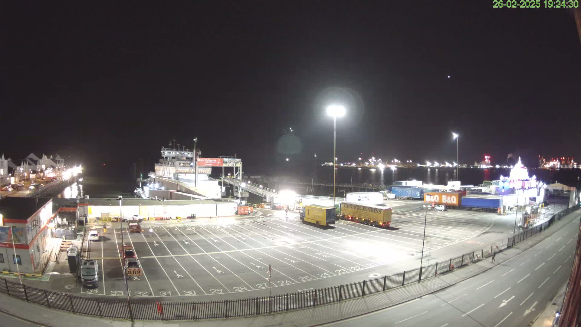 A nighttime, brightly lit view of a ferry terminal with several trucks parked in an empty lot, a ferry docked, and a body of water visible in the background.