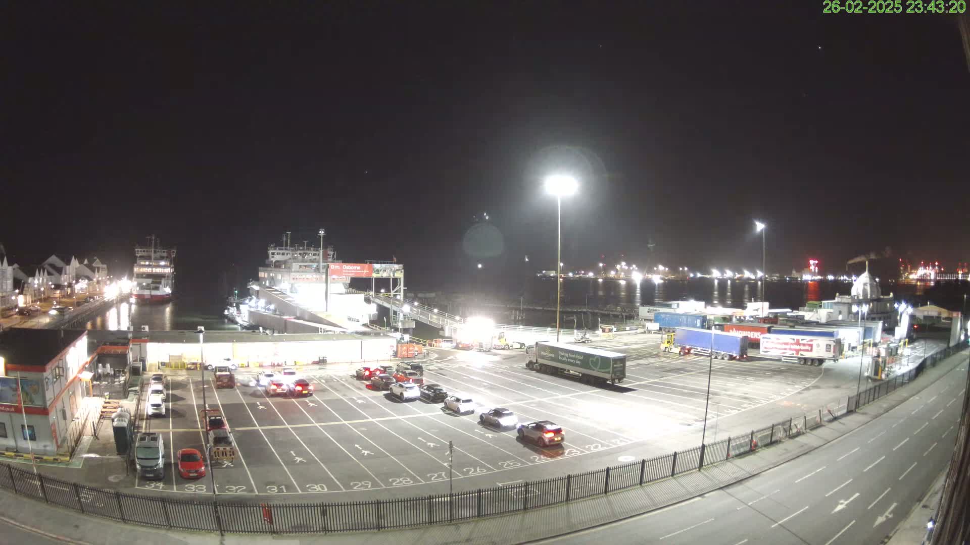 A nighttime view of a ferry terminal with several cars parked in a lot, a ferry docked, and numerous trucks in the background.