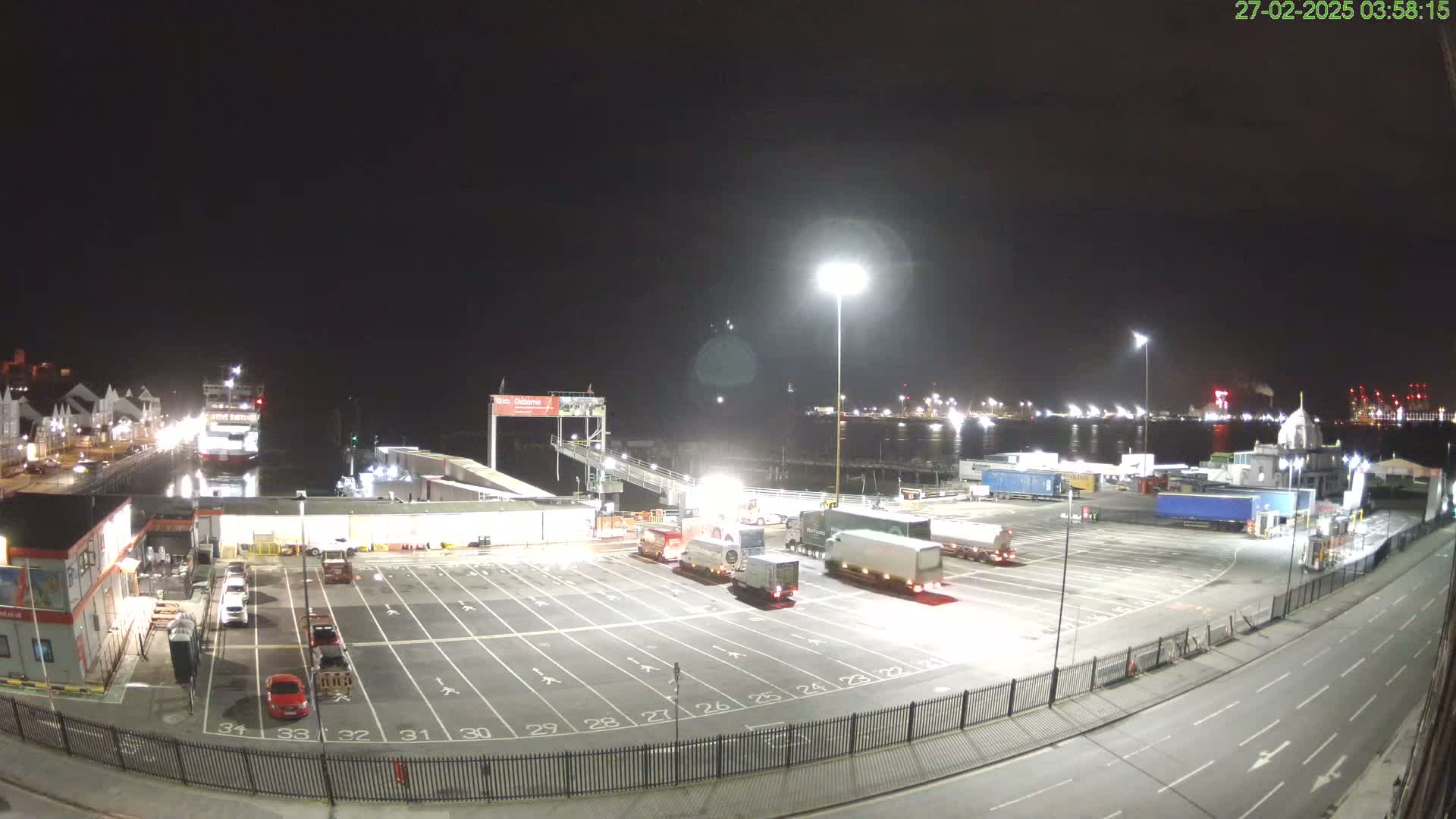 A nighttime view of a ferry terminal with several trucks parked in a lot, a ferry docked, and a body of water in the background under a clear, dark sky.