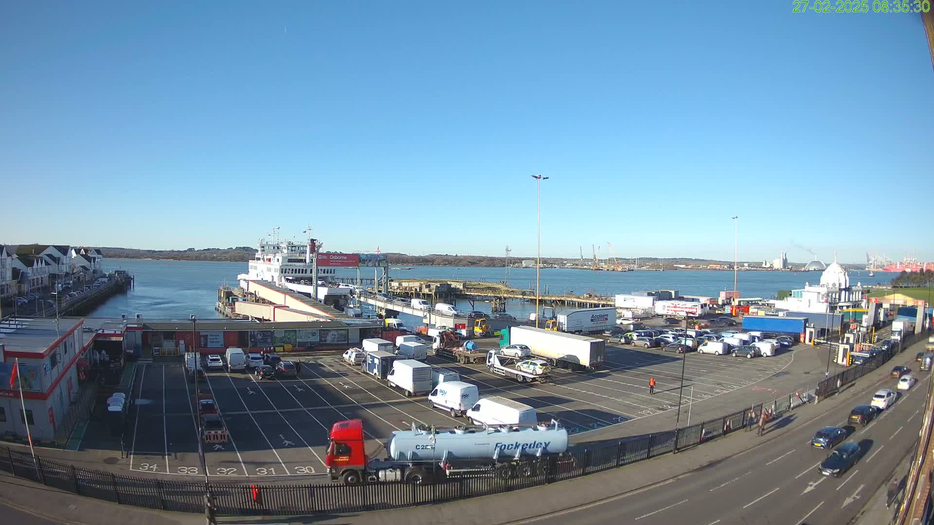 A ferry is docked at a port on a sunny day, with numerous vehicles parked in the surrounding lot and several more driving on a nearby road.