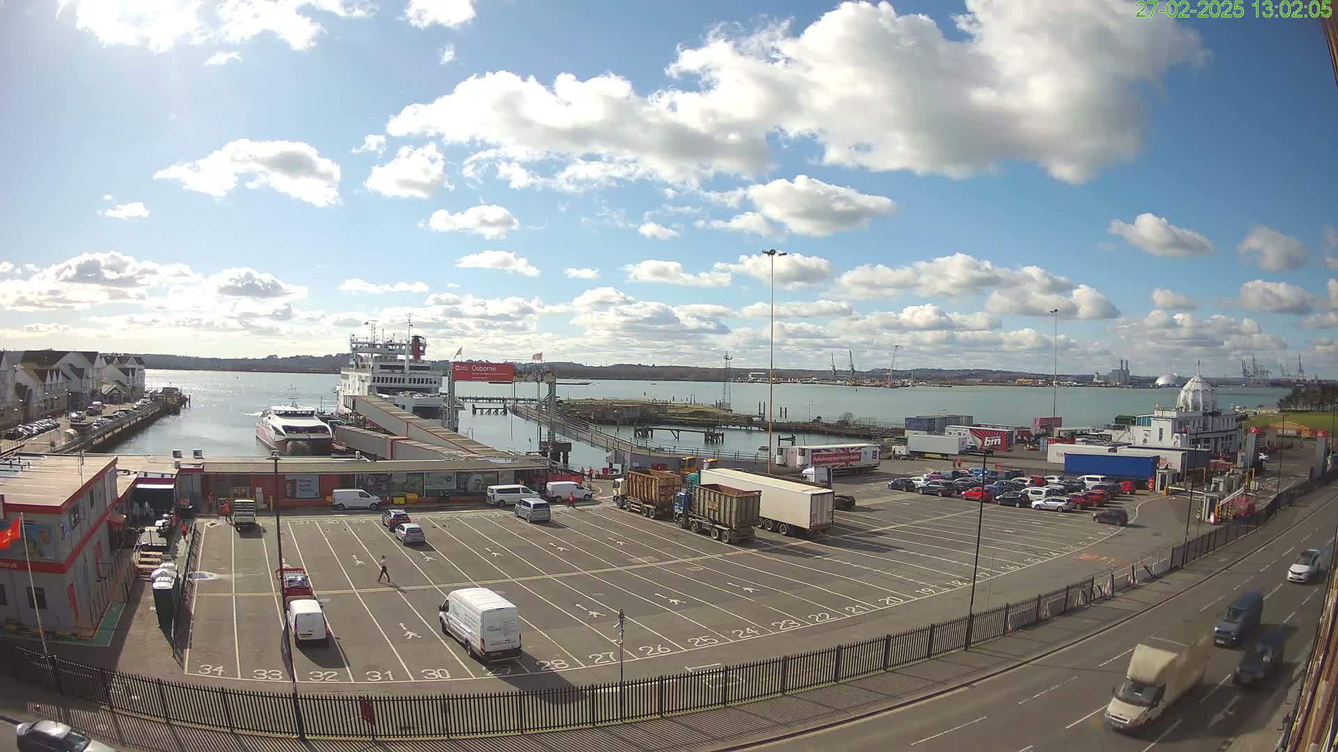 A ferry is docked at a port under a partly sunny sky with scattered clouds; vehicles are parked in a lot nearby, and more are on the road beyond.