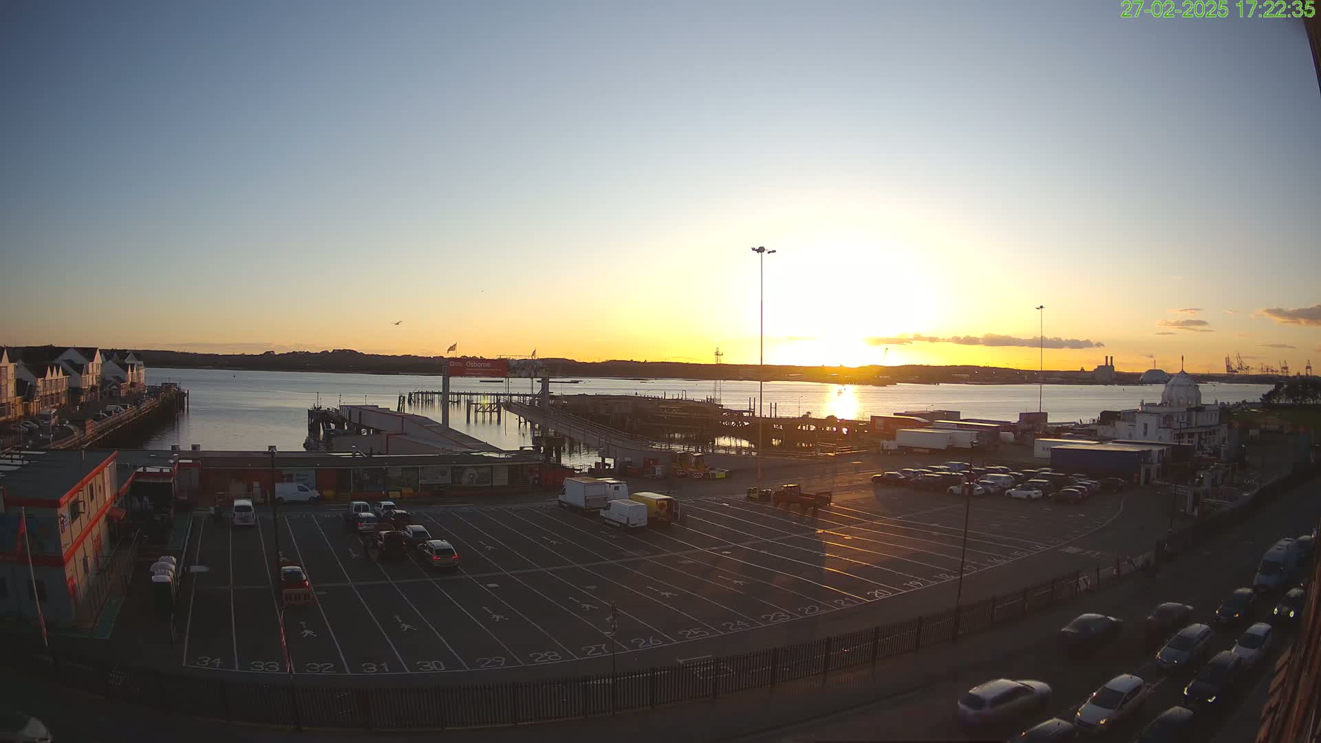 A sunset over a calm body of water, showing a ferry terminal with a parking lot filled with cars, and buildings along the waterfront.