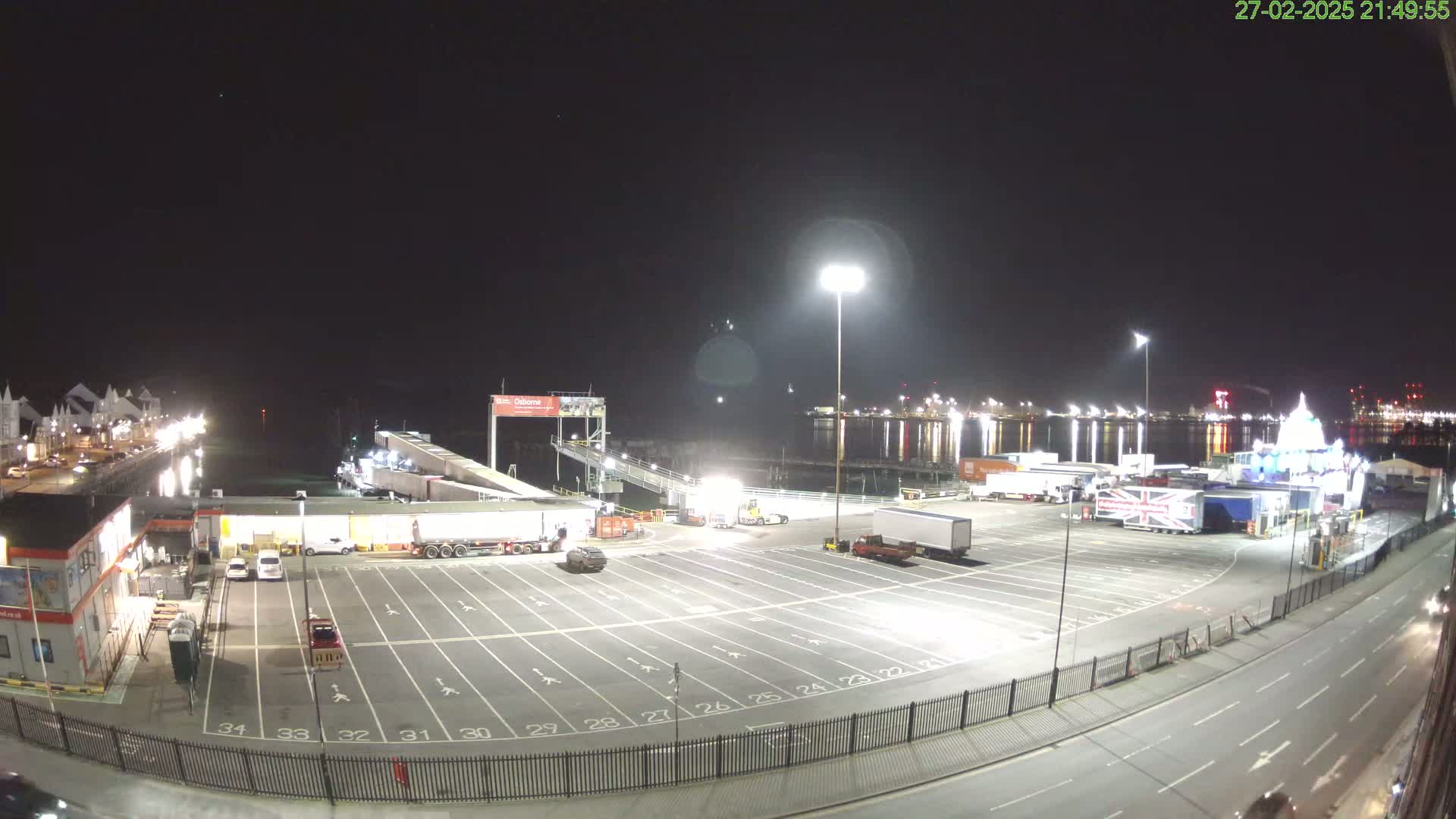 A nighttime view of a ferry terminal with several trucks and cars parked in a large lot, next to a body of water reflecting lights.