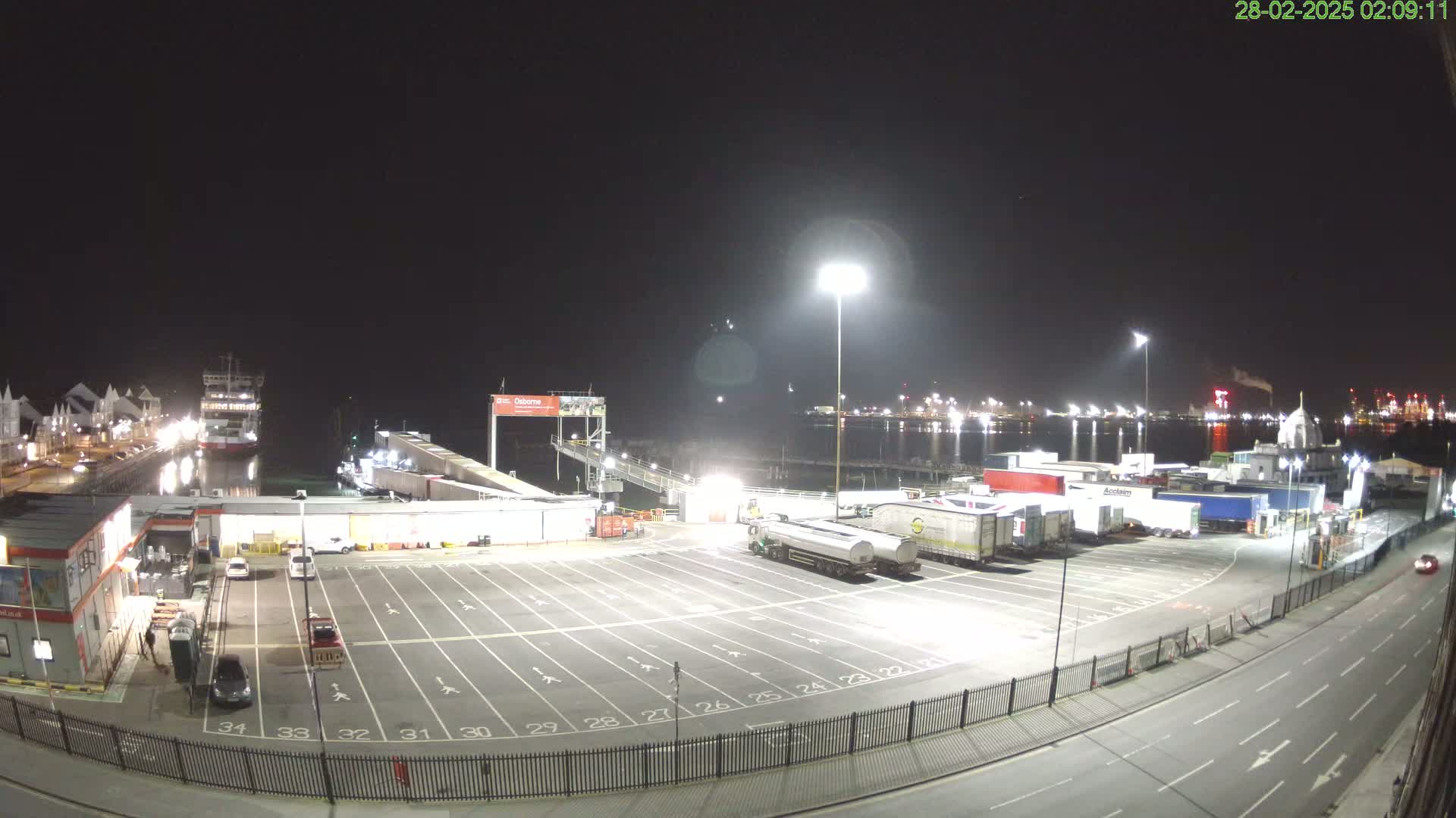 A nighttime view of a ferry terminal with a ferry docked, numerous trucks parked in a lot, and a roadway visible in the foreground under clear skies.