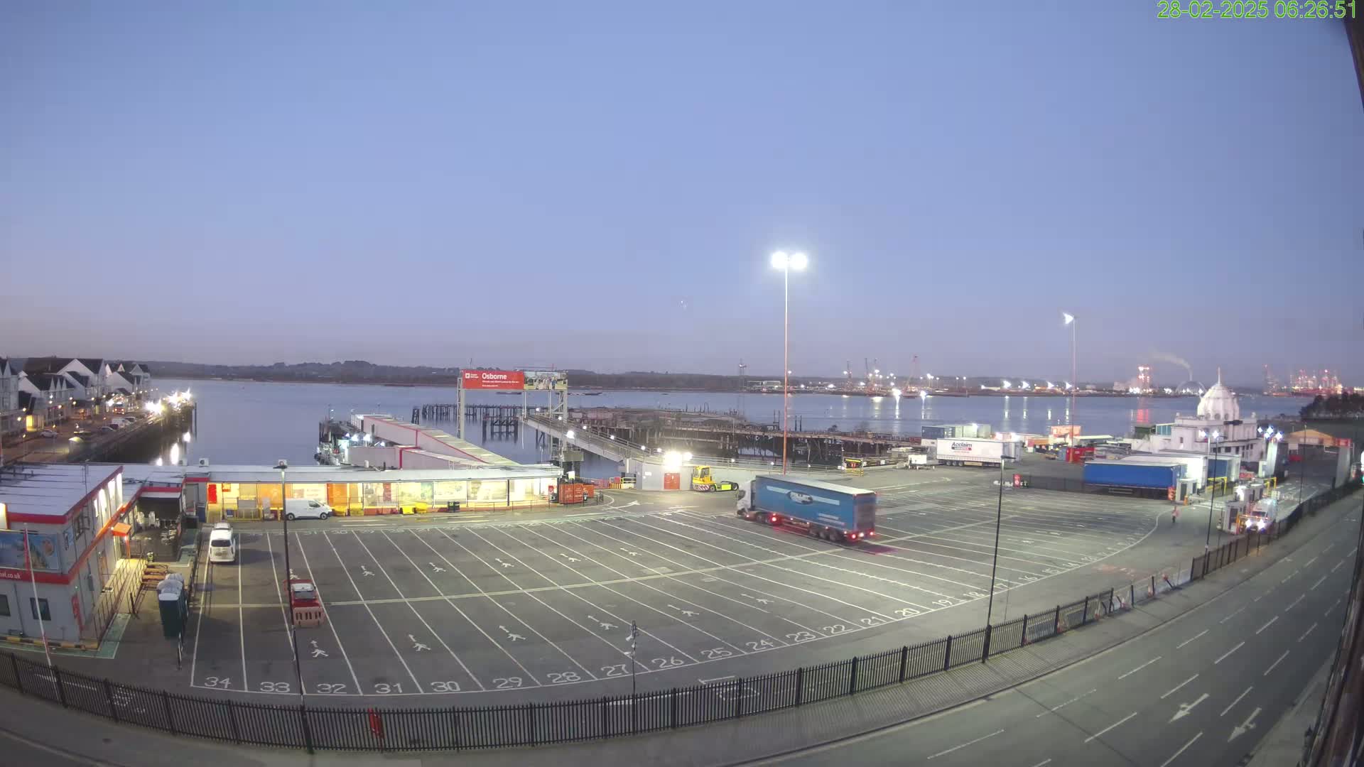 A large empty parking lot at a ferry terminal overlooks a calm body of water at dawn.