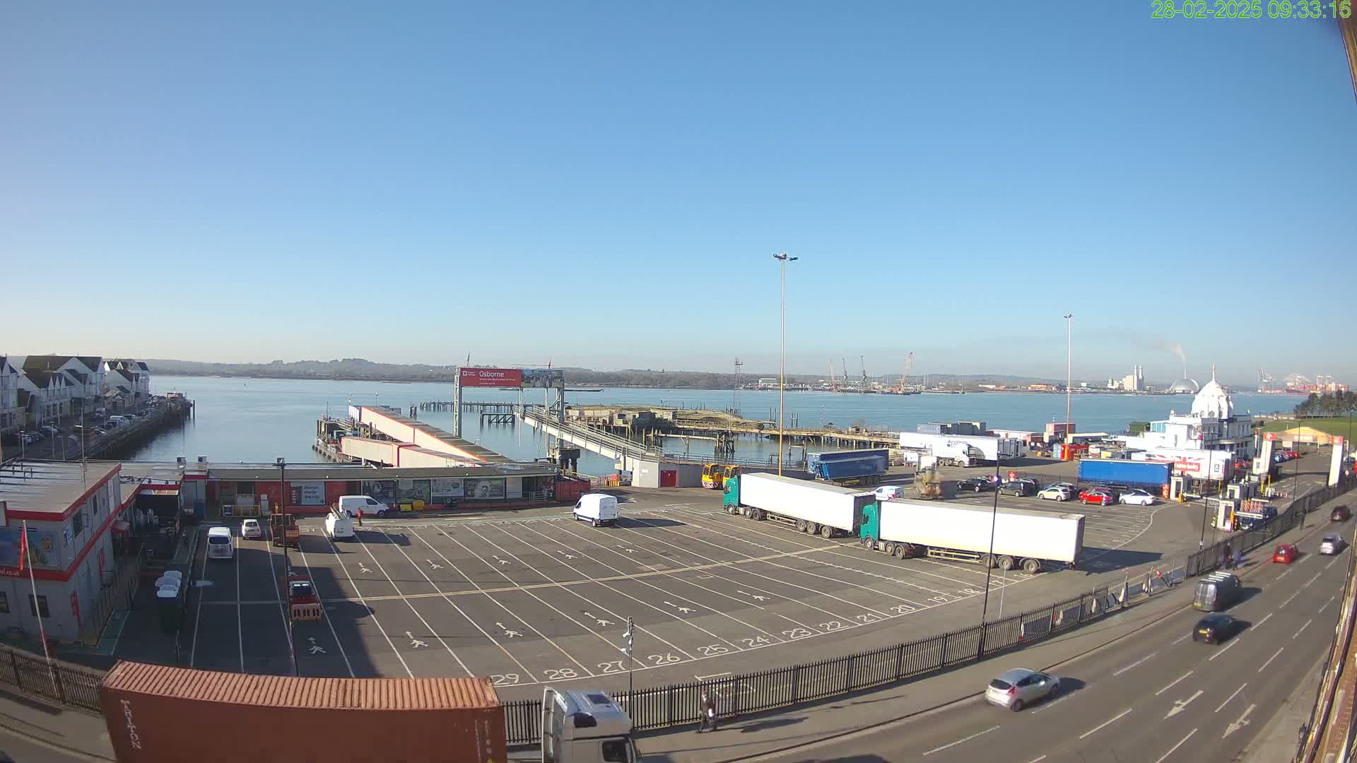 A ferry terminal with a large empty parking lot for trucks and cars, under a clear blue sky.