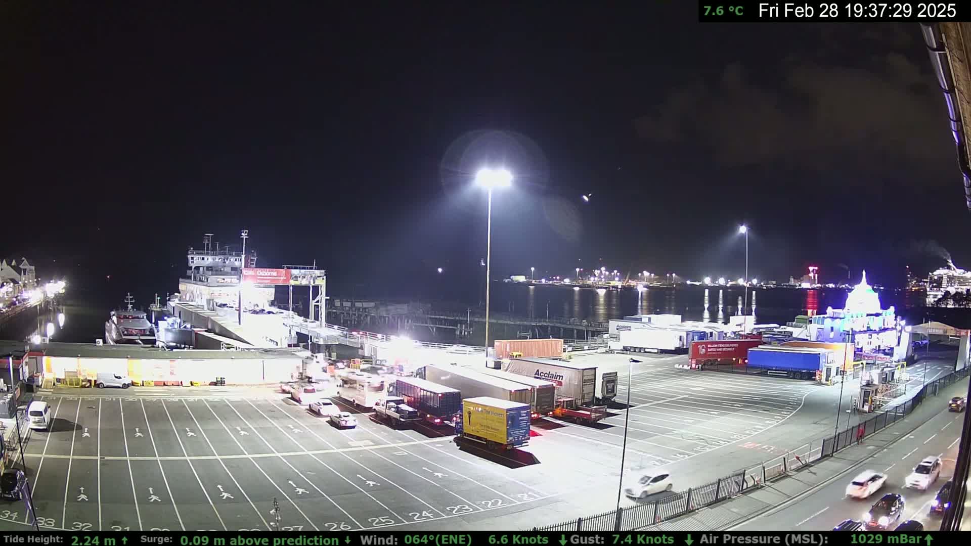 A ferry is docked at a port at night, with several trucks and cars parked in the surrounding lot under bright lights, and a calm body of water visible in the background.