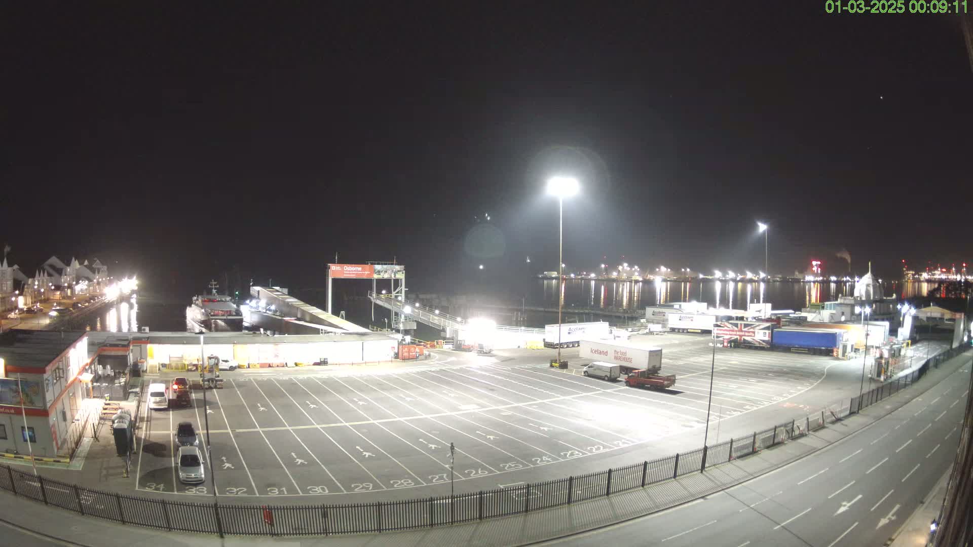 A nighttime view of a ferry terminal with a boat docked, several parked cars and trucks, and a large empty parking lot, under a clear sky.