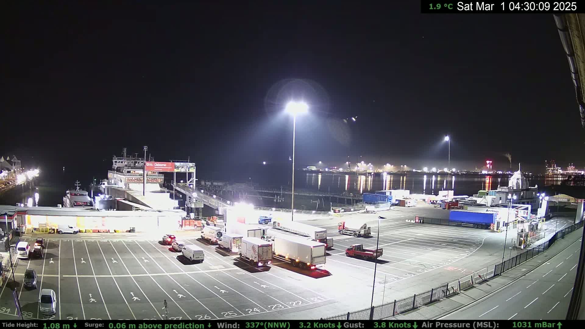 A nighttime view of a ferry terminal with several vehicles parked in the lot, a ferry docked, and calm water reflecting lights in the background under a clear sky.