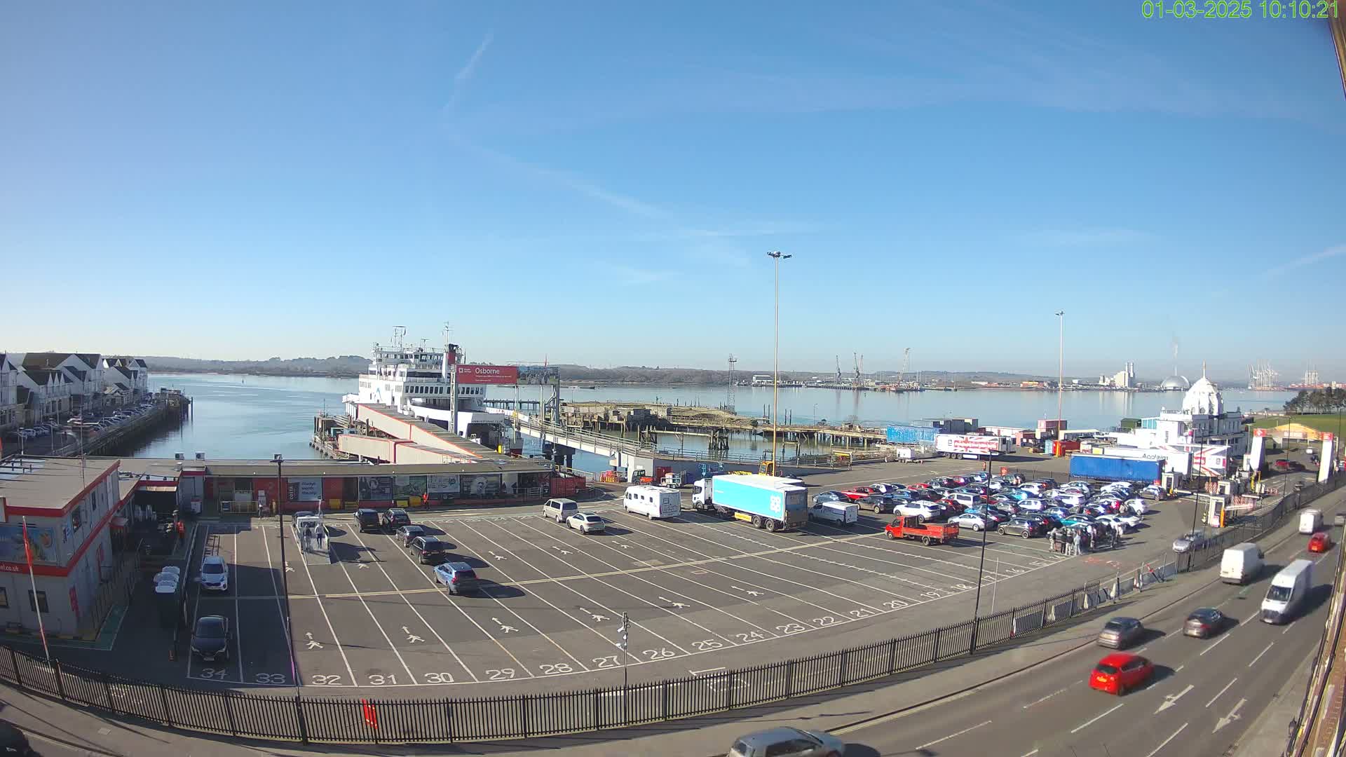 A ferry is docked at a port on a sunny day, with numerous cars parked in a lot nearby and traffic on the road beyond.