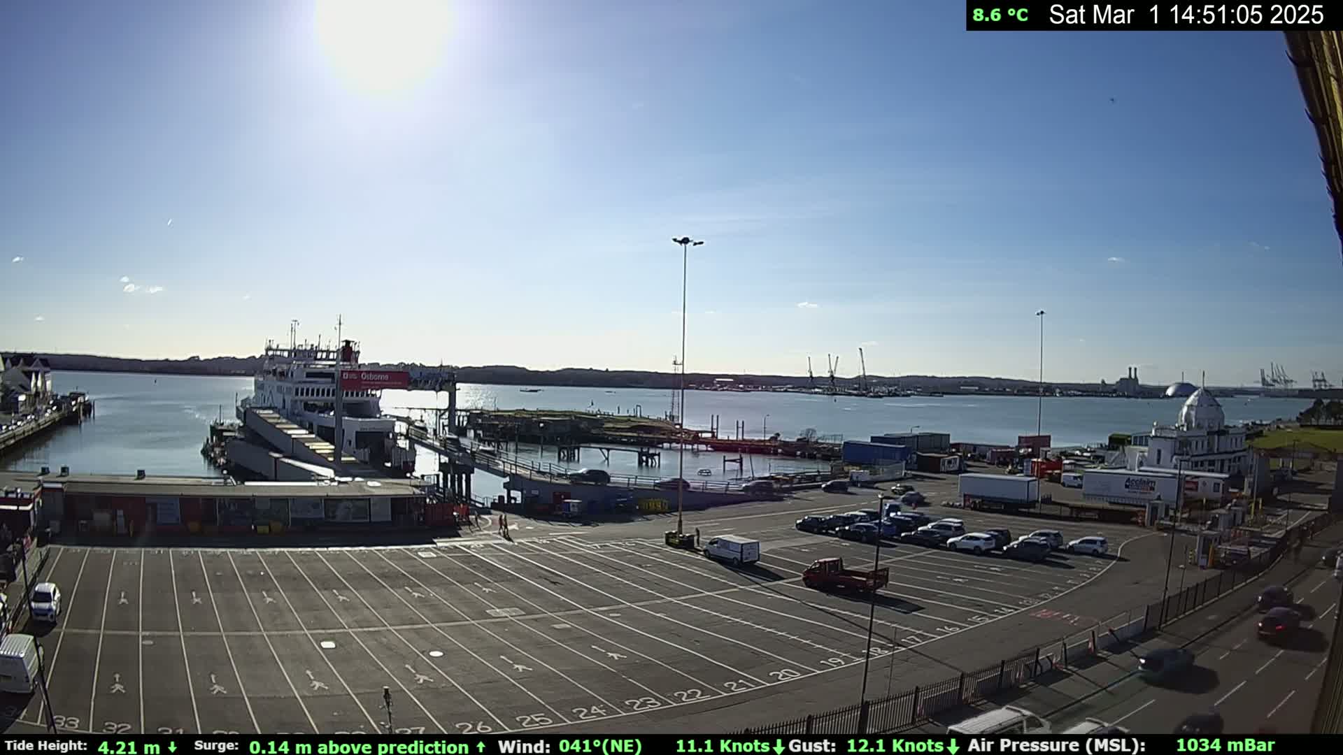 A ferry is docked at a port on a sunny day with a calm body of water, and a large empty parking lot is visible with several cars parked.