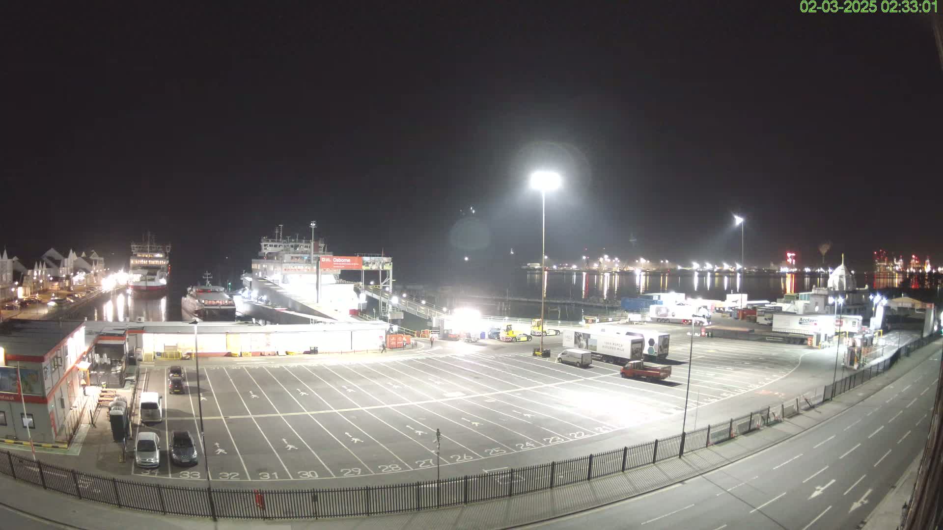 A nighttime view of a ferry terminal with several ships docked, a large empty parking lot, and numerous trucks and cars present.
