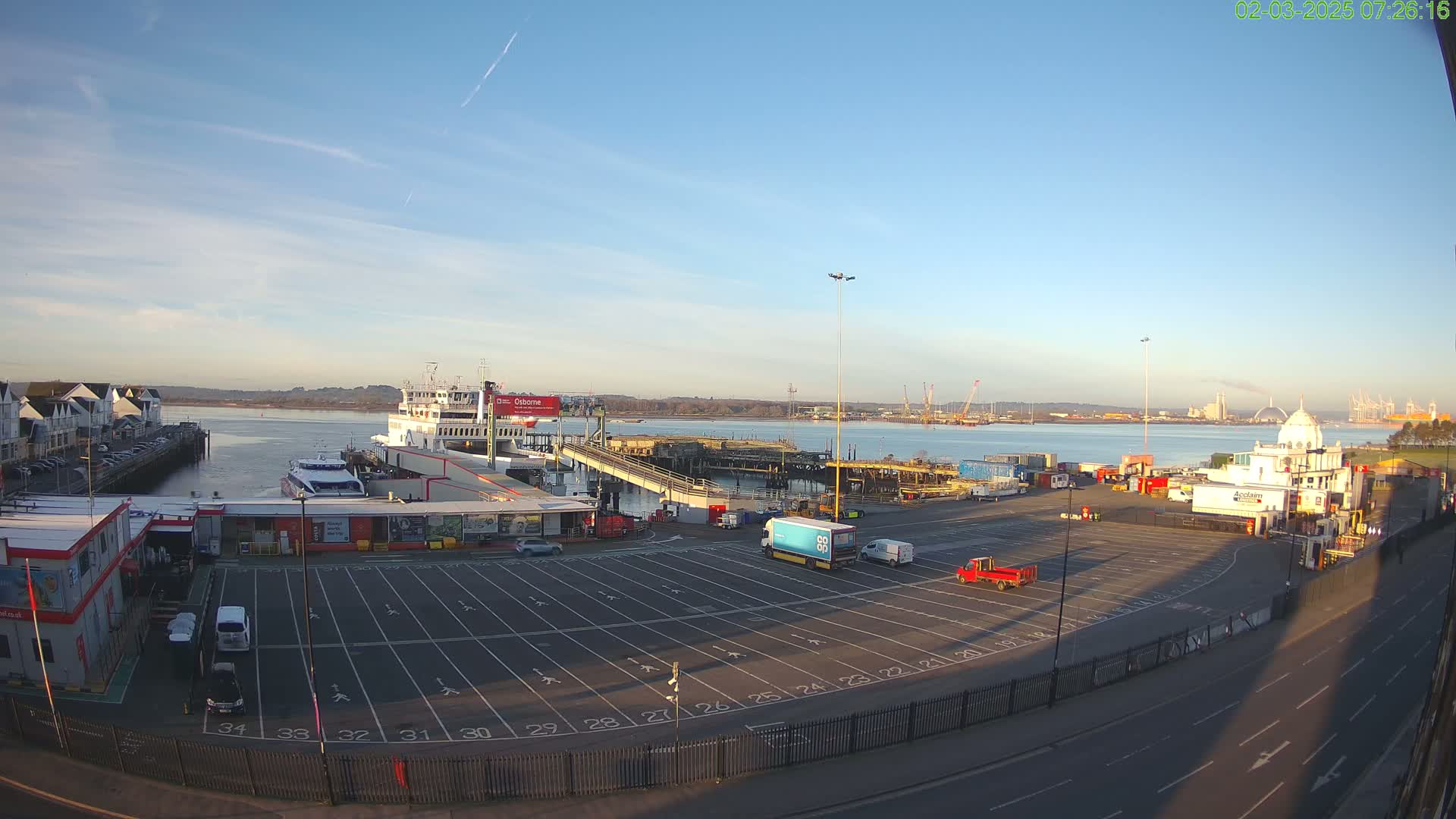 A ferry is docked at a terminal on a sunny day, with a mostly clear sky and calm water in the background.