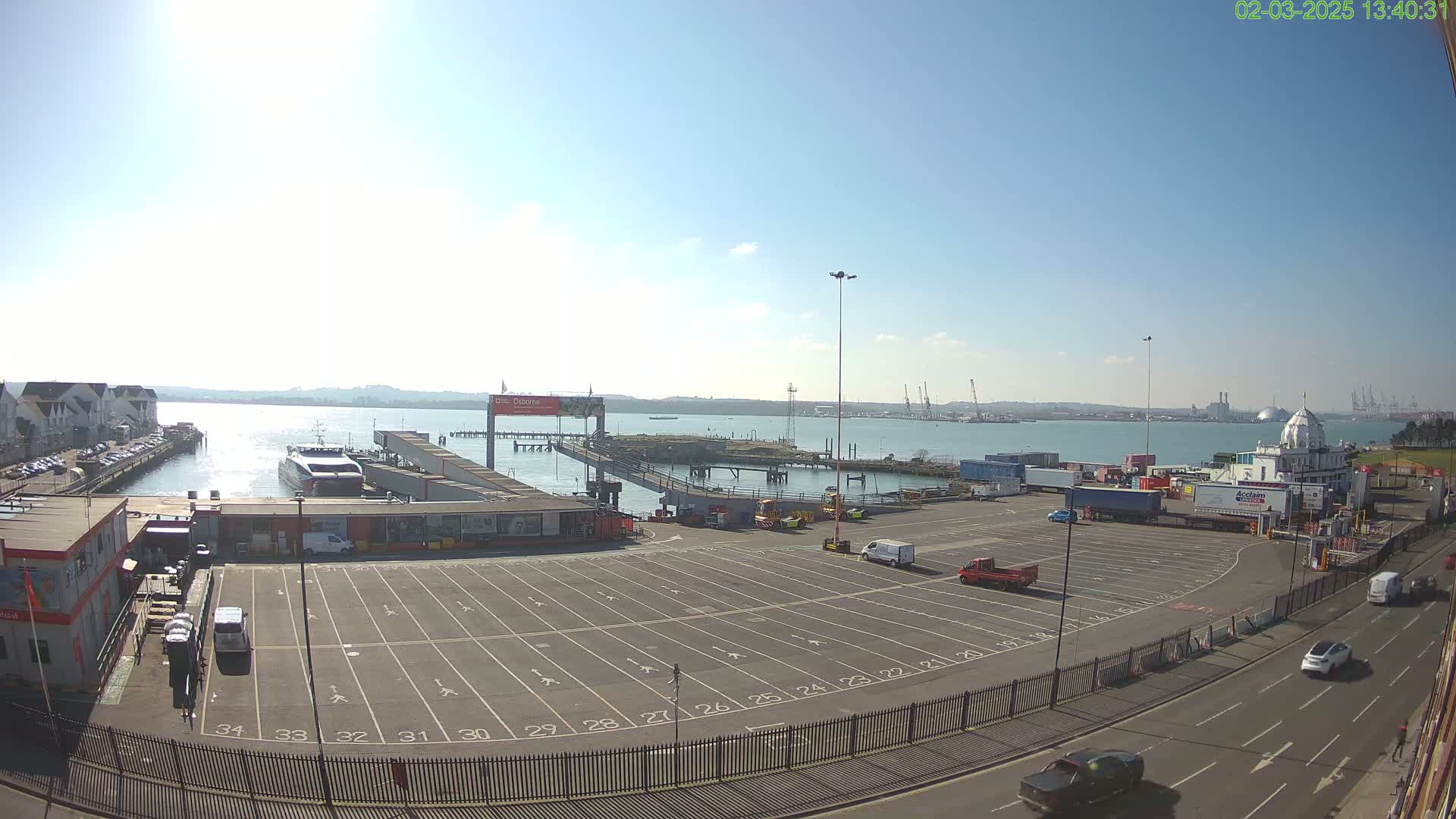 A sunny day at a ferry terminal shows a large empty parking lot, a ferry docked at the pier, and several vehicles in the surrounding area.