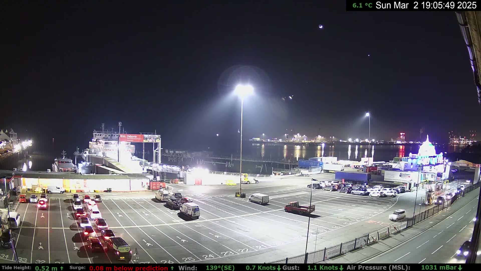 A ferry is docked at a terminal at night under a clear sky with a few stars visible;  cars are parked in a lot, and several vehicles are waiting in lines to board.