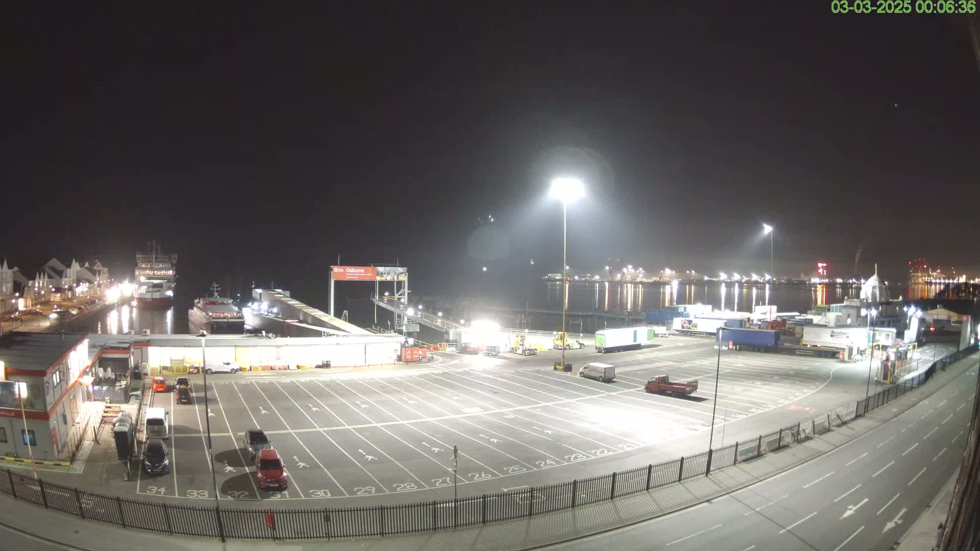 A nighttime view of a port with several ships docked, a large empty parking lot, and various trucks and vehicles.