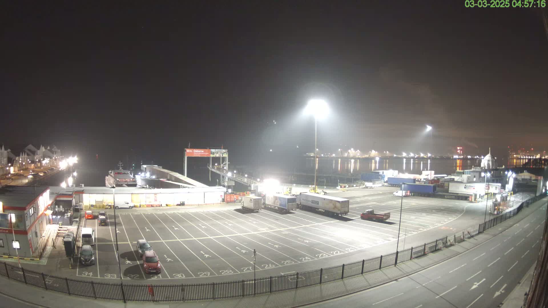 A nighttime view of a ferry terminal with a mostly empty parking lot for trucks and cars, a ferry docked, and bright lights illuminating the area.