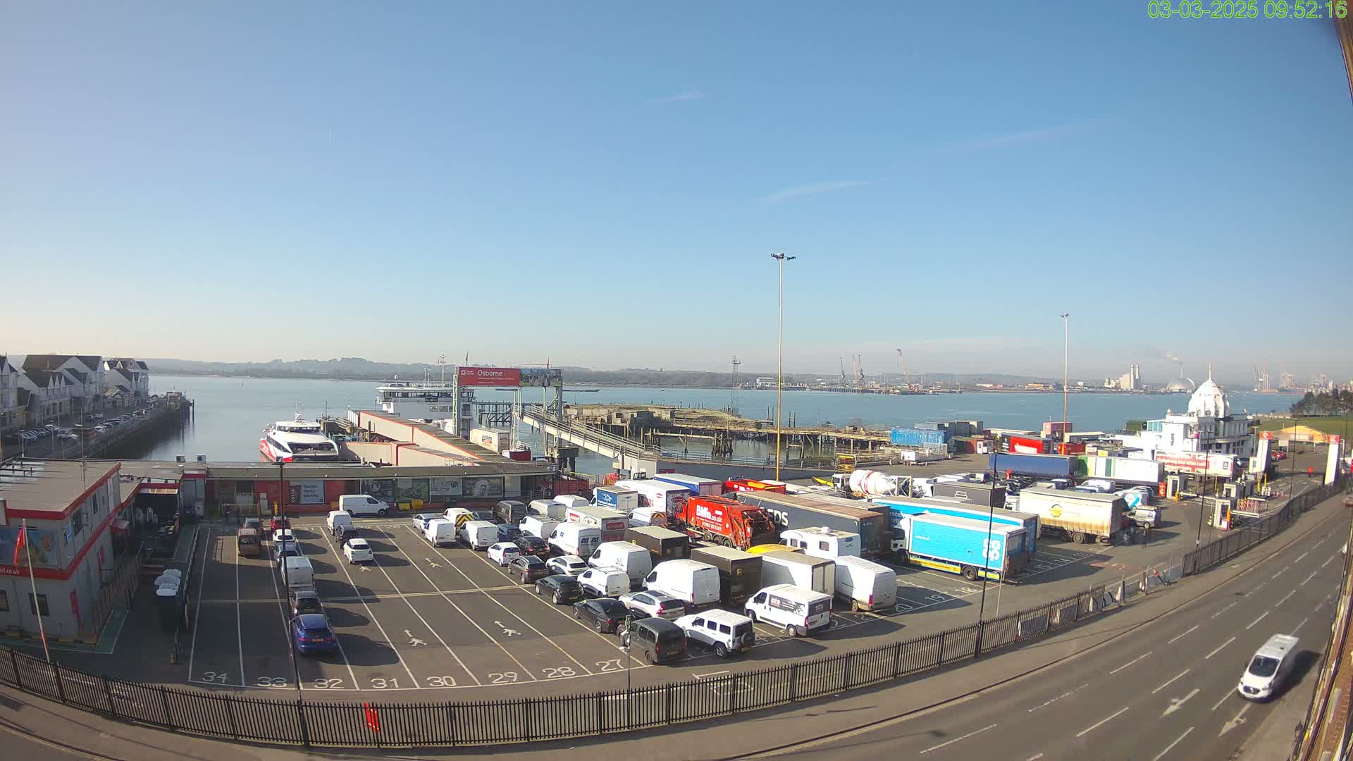 A ferry terminal on a sunny day shows a ferry docked, numerous vehicles parked in a lot, and a body of water with a distant shoreline.