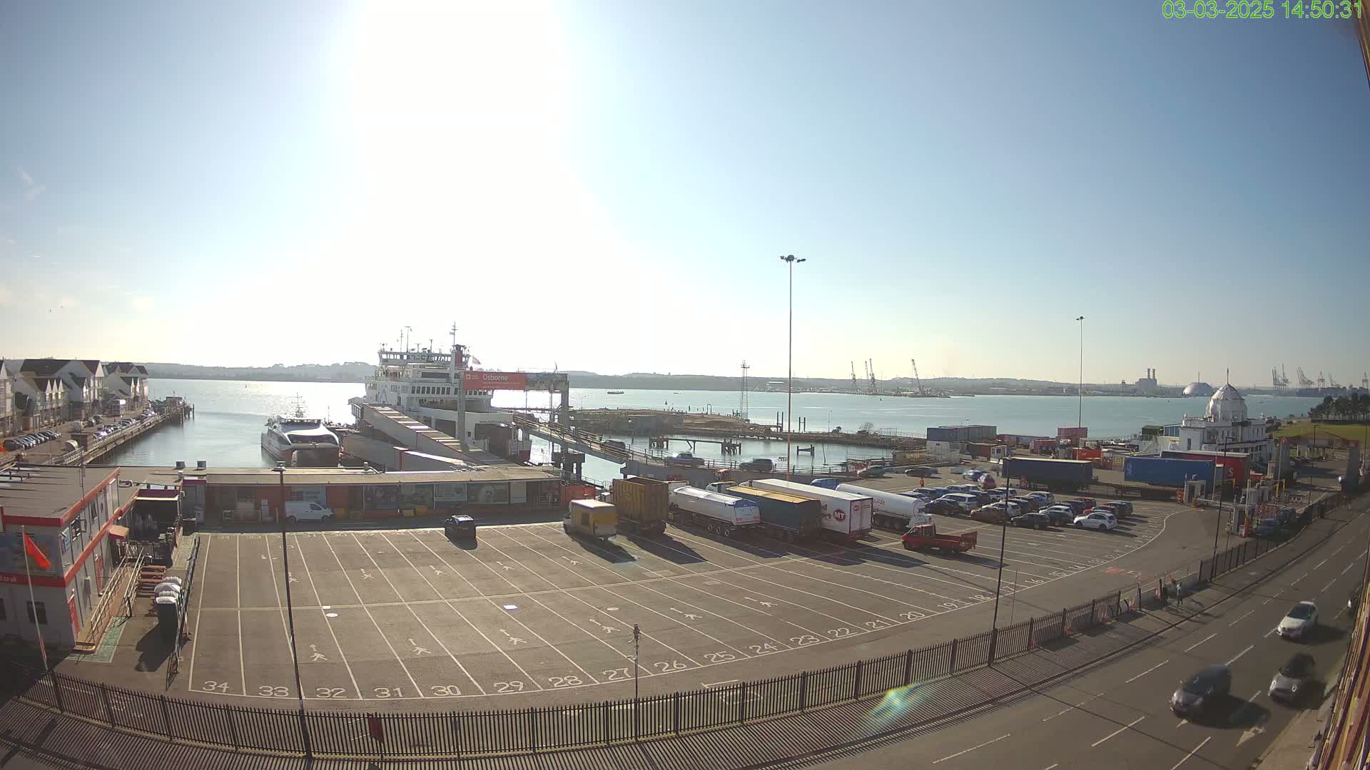 A ferry is docked at a port on a sunny day, with numerous vehicles parked in a lot nearby and a body of water visible in the background.