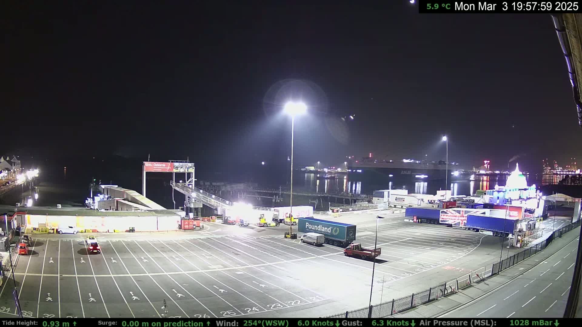 A nighttime view of a ferry terminal with several trucks and cars parked in a large, empty lot, under a clear sky.