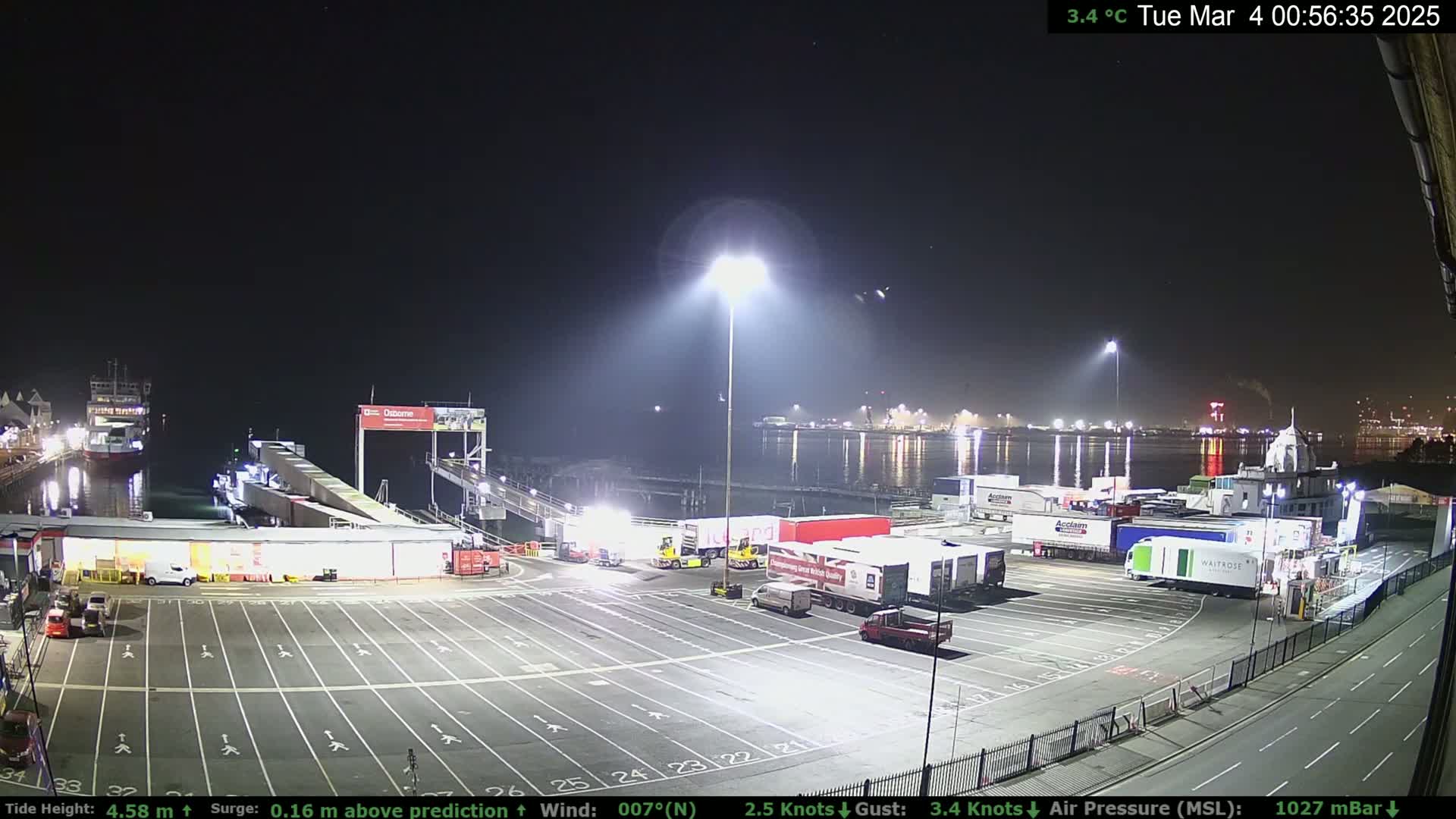 A nighttime view of a ferry terminal with numerous trucks parked in a large lot, a ferry docked, and calm water visible in the background under a clear sky.