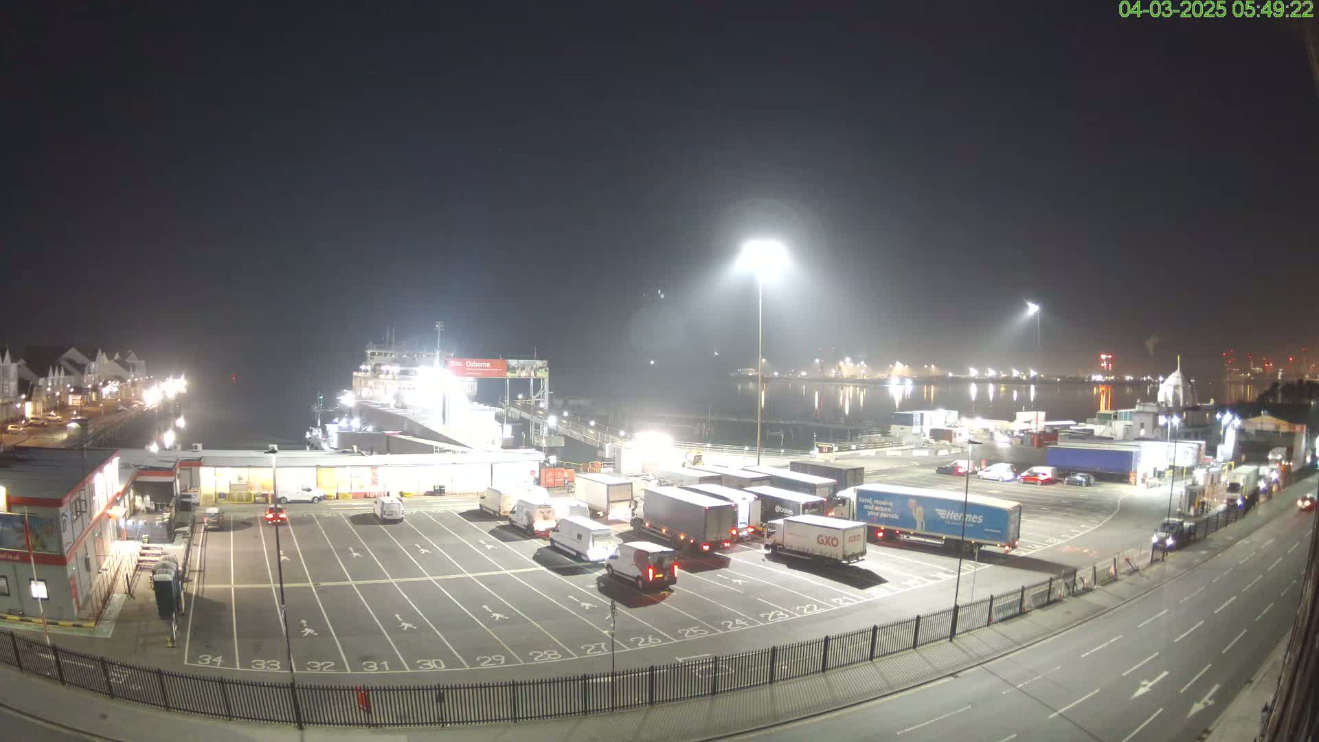 A nighttime view of a ferry terminal with numerous trucks and cars parked in a lot, a ferry docked, and calm water in the background under a hazy sky.