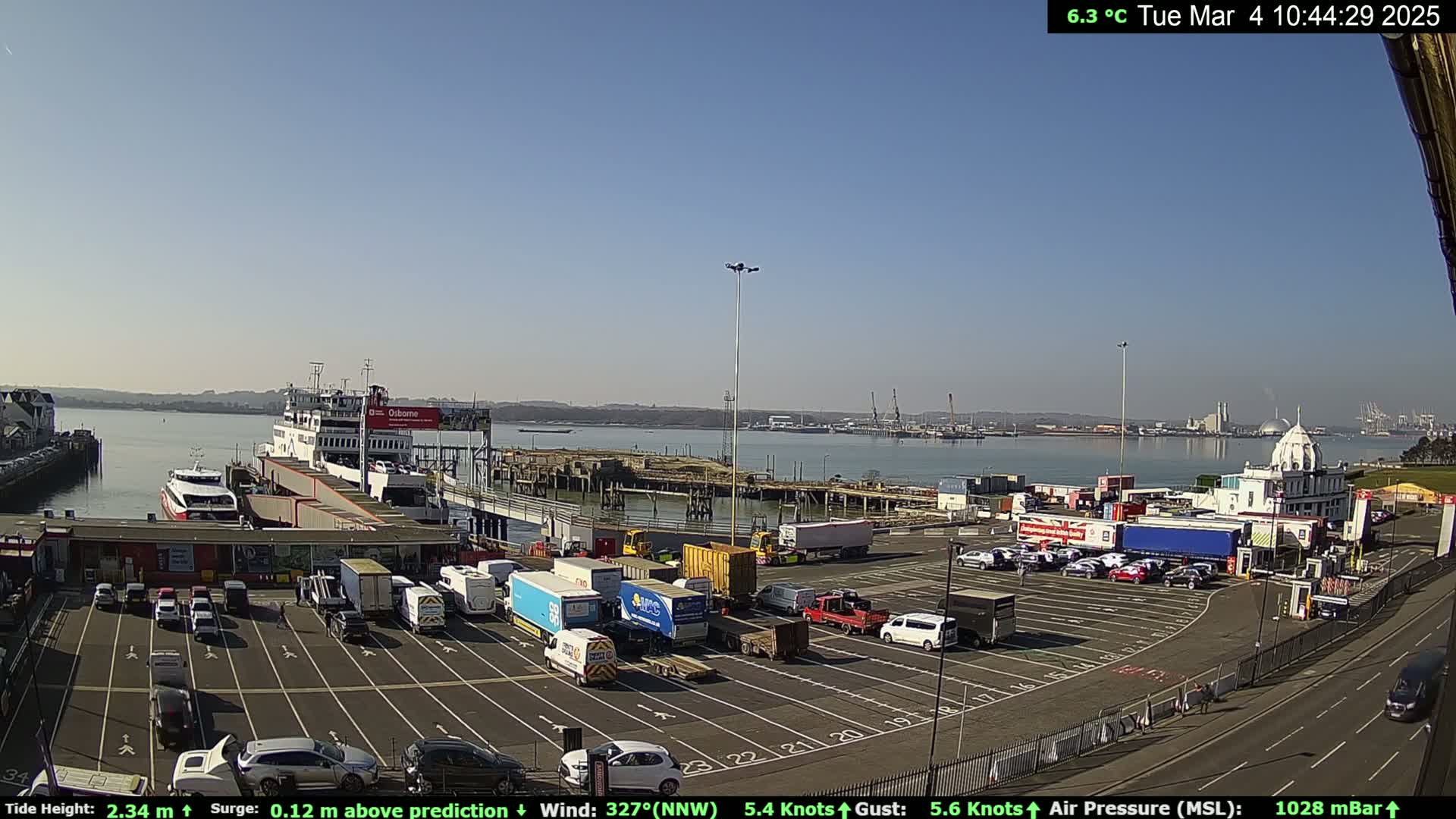 A ferry is docked at a terminal with numerous vehicles parked in a lot, under a clear sunny sky.