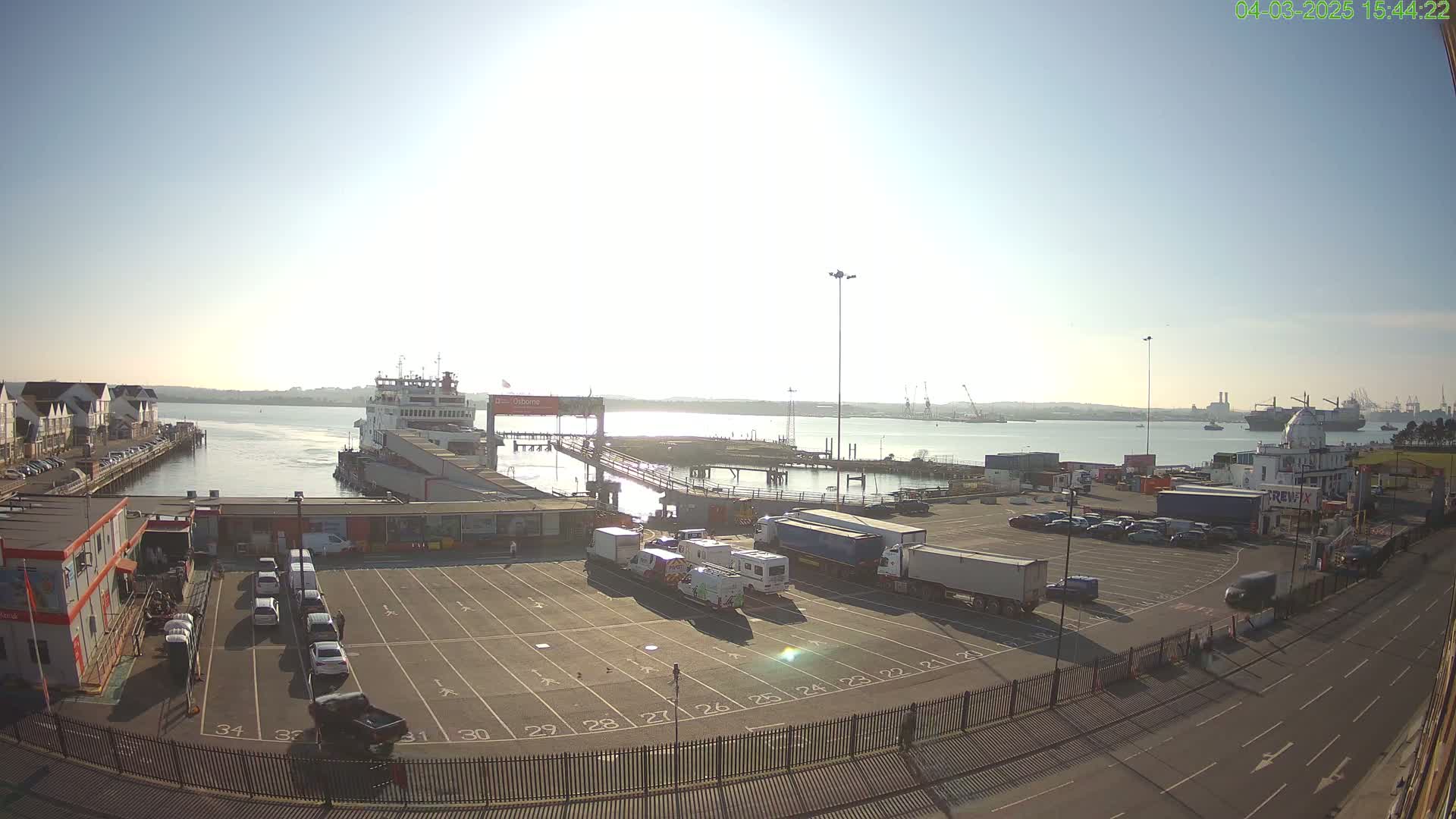 A ferry is docked at a port on a sunny day, with a large empty parking lot filled with a few cars and trucks in front of it, and a body of water with other ships in the distance.