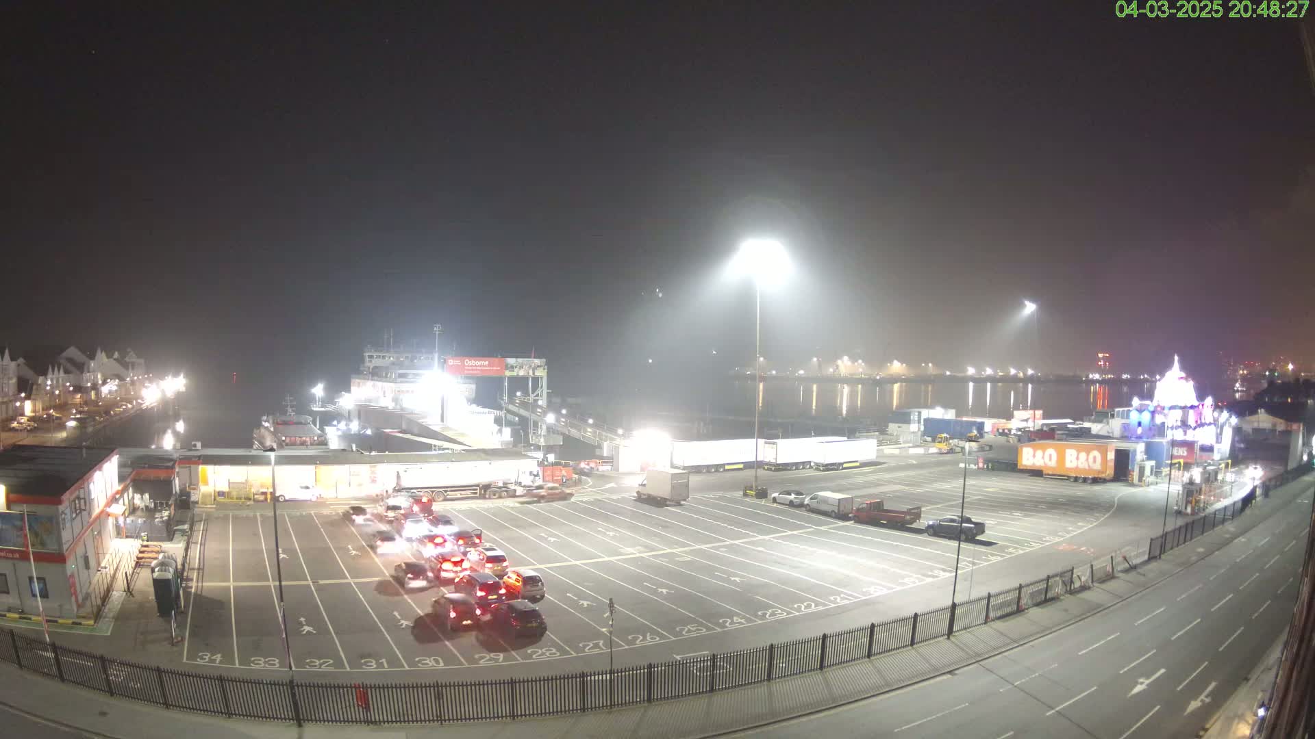 A nighttime, hazy view of a ferry terminal with numerous vehicles parked and waiting, along with a few moving on the road.