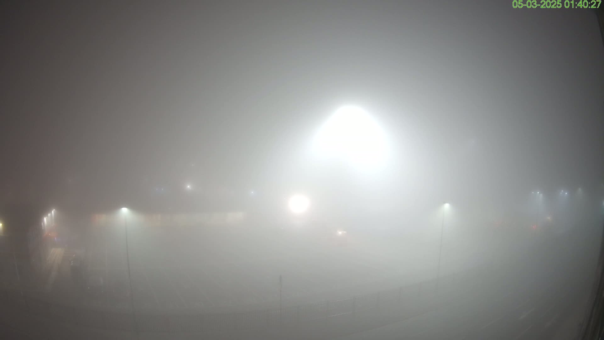 A thick fog obscures a dimly lit, empty parking lot or field at night, with several bright lights barely visible through the haze.