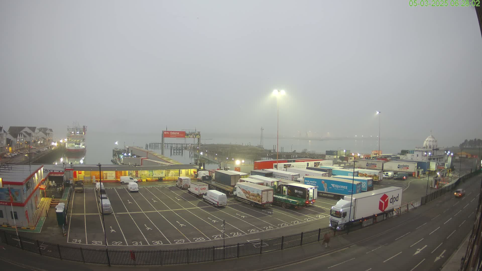 A foggy morning scene at a port shows a ferry docked, numerous delivery trucks parked in a lot, and a roadway.